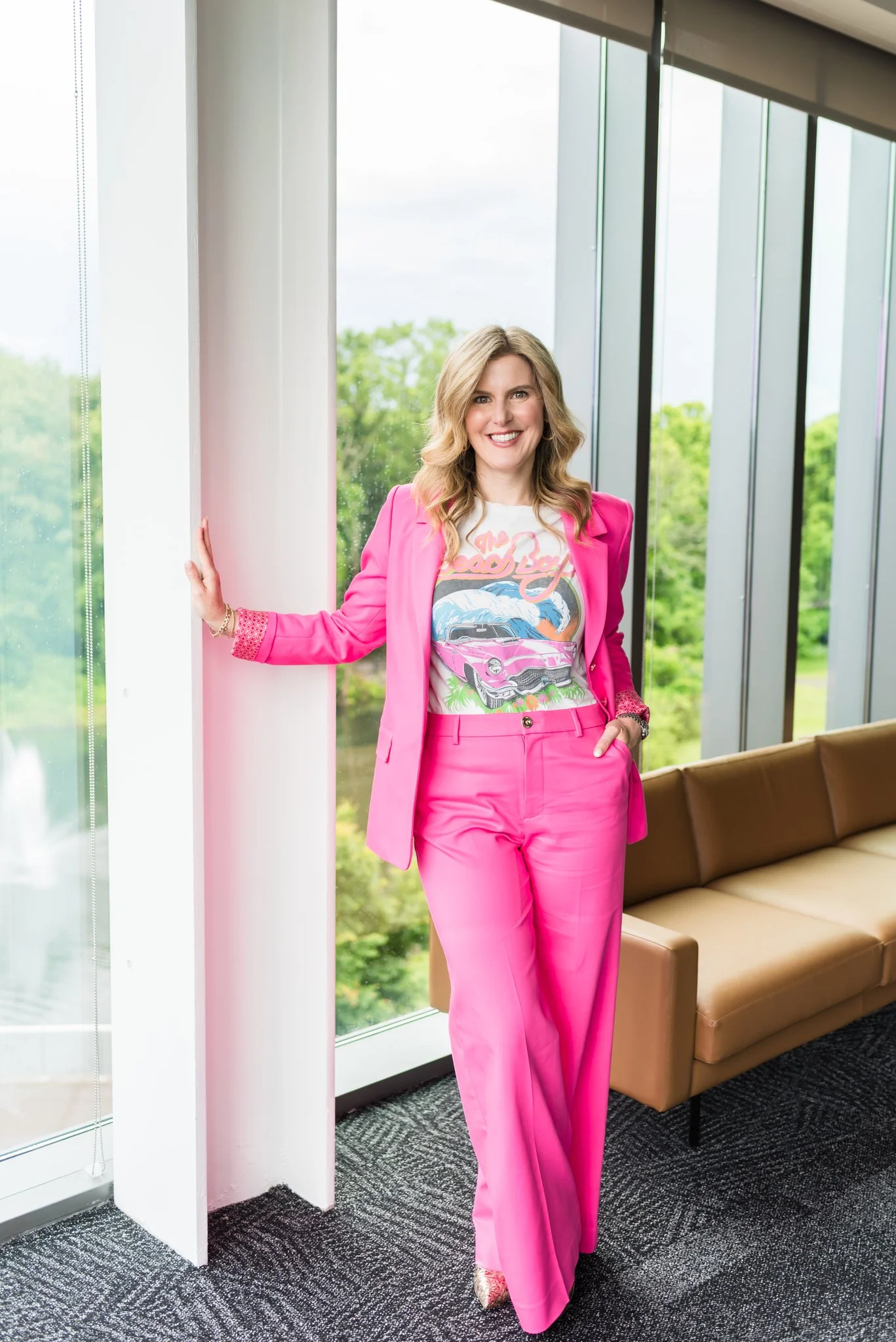 Woman in a pink suit, pink pants and blazer, with a graphic t-shirt, standing next to a large window, smiling, with a green outdoor view.