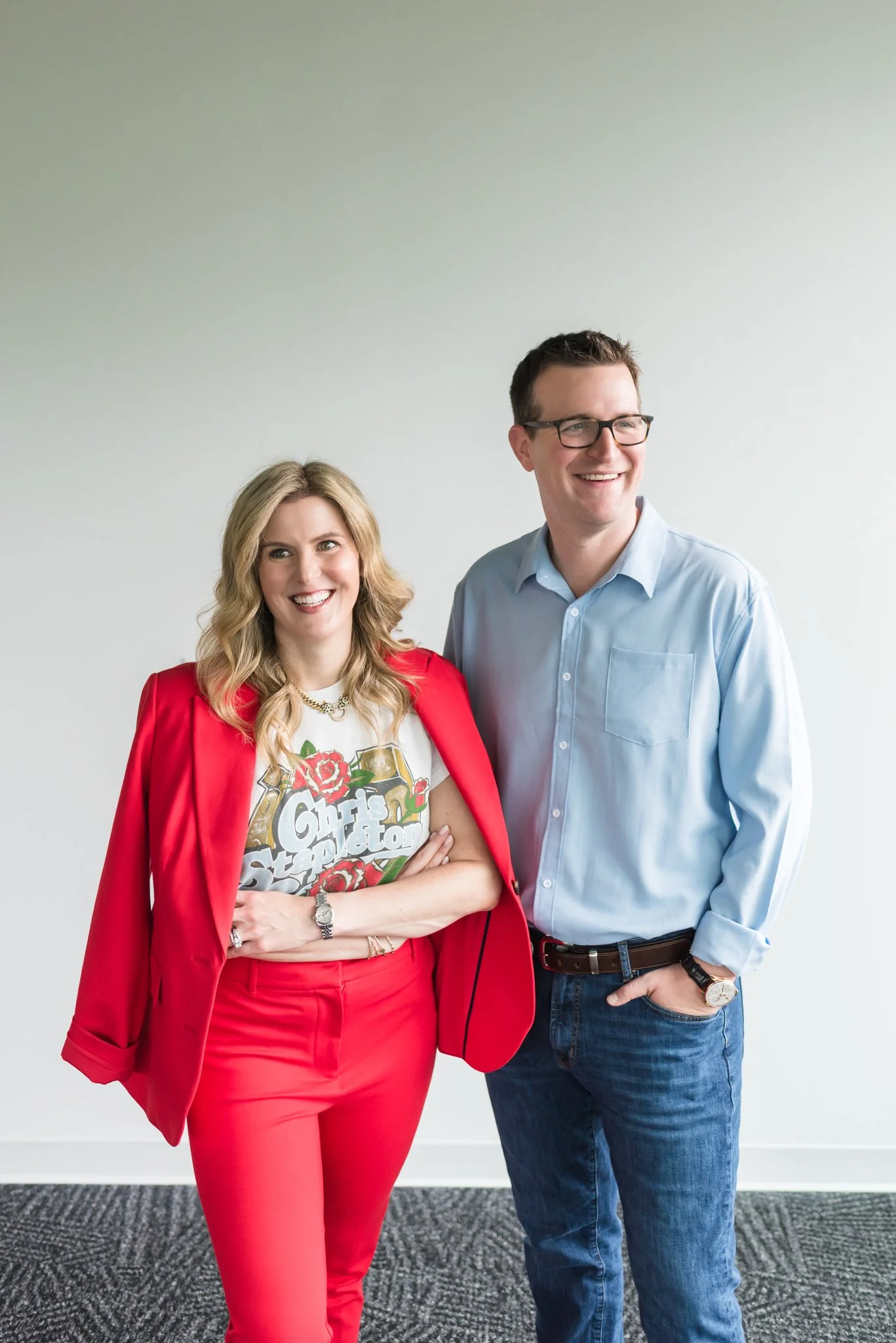 A woman in a red suit with a graphic t-shirt and a man in a light blue shirt and jeans standing together and smiling against a plain white wall.