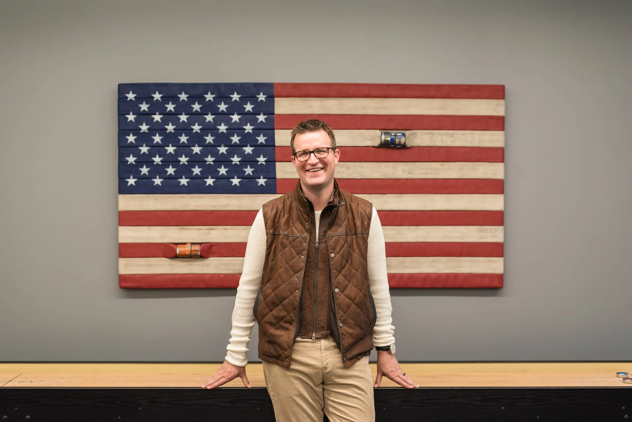 Man wearing glasses, a brown vest over a cream sweater, smiling, standing in front of a large American flag made of wooden planks on a gray wall.