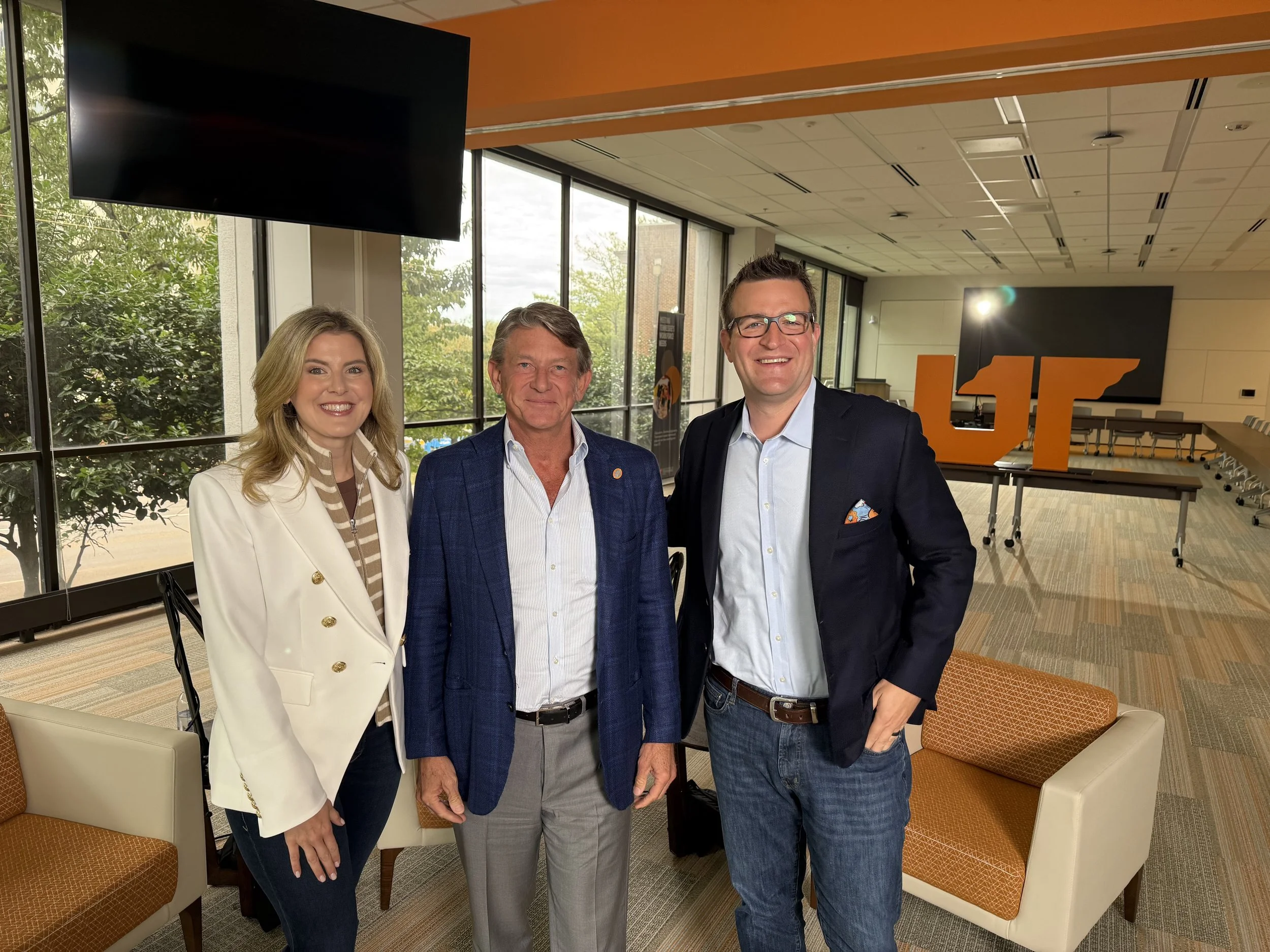 Three people standing in a conference room with large windows, a table, and a large orange 'UF' sign in the background.