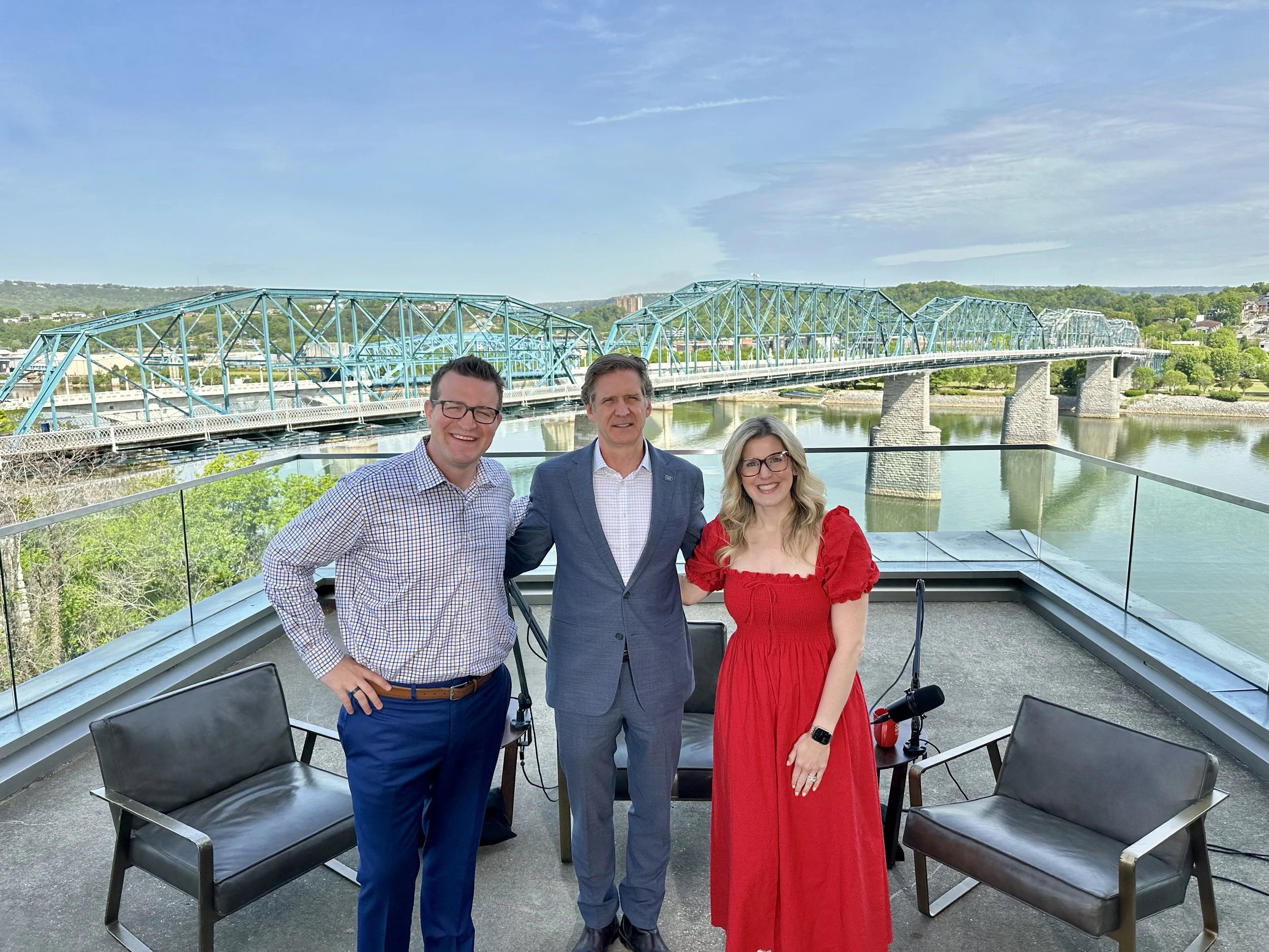 Three people standing on a rooftop balcony, with a river and a blue bridge in the background, during daytime.