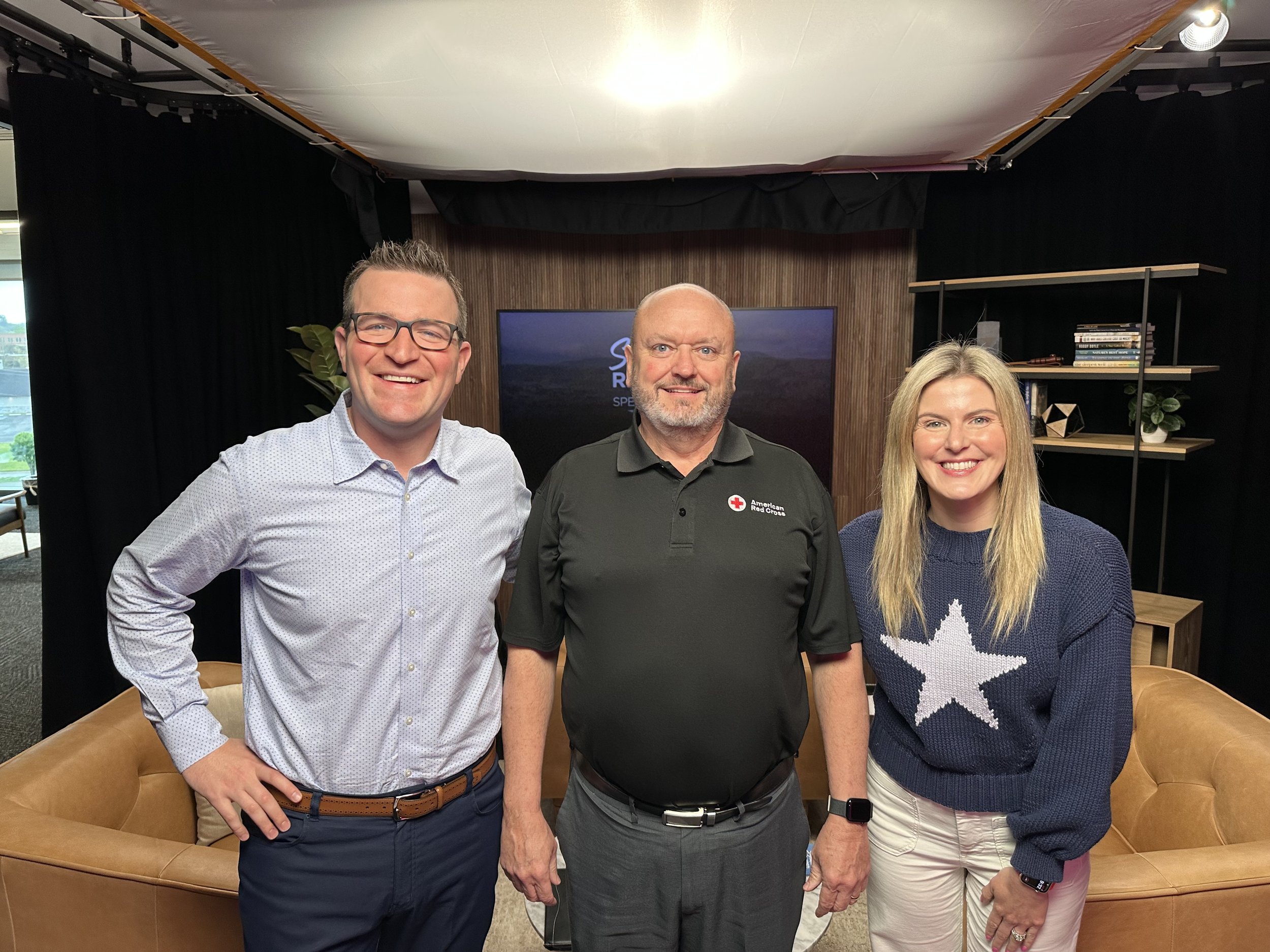 Three smiling people standing in a studio set with a sofa, bookshelf, and TV in the background.