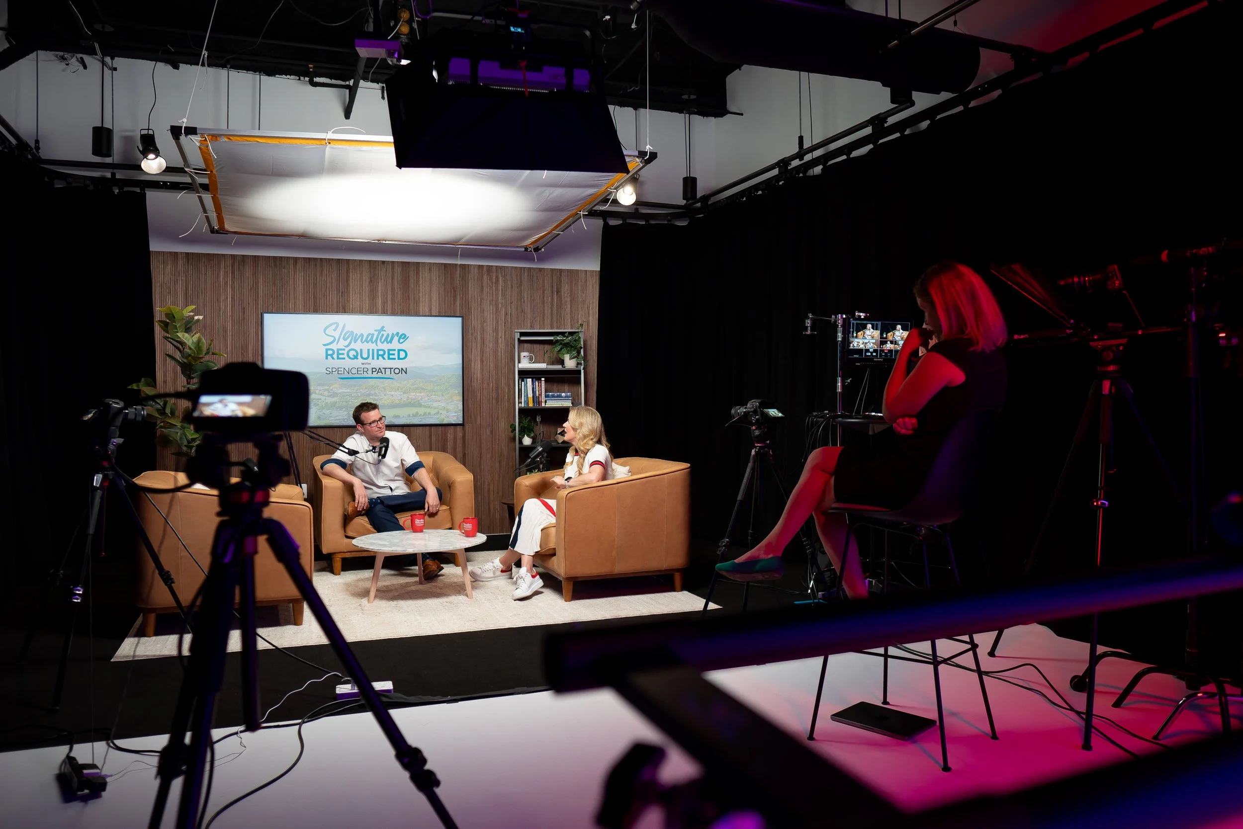 Two people seated on brown sofas having a discussion in a studio, with a woman sitting on a chair recording them and camera equipment around them.