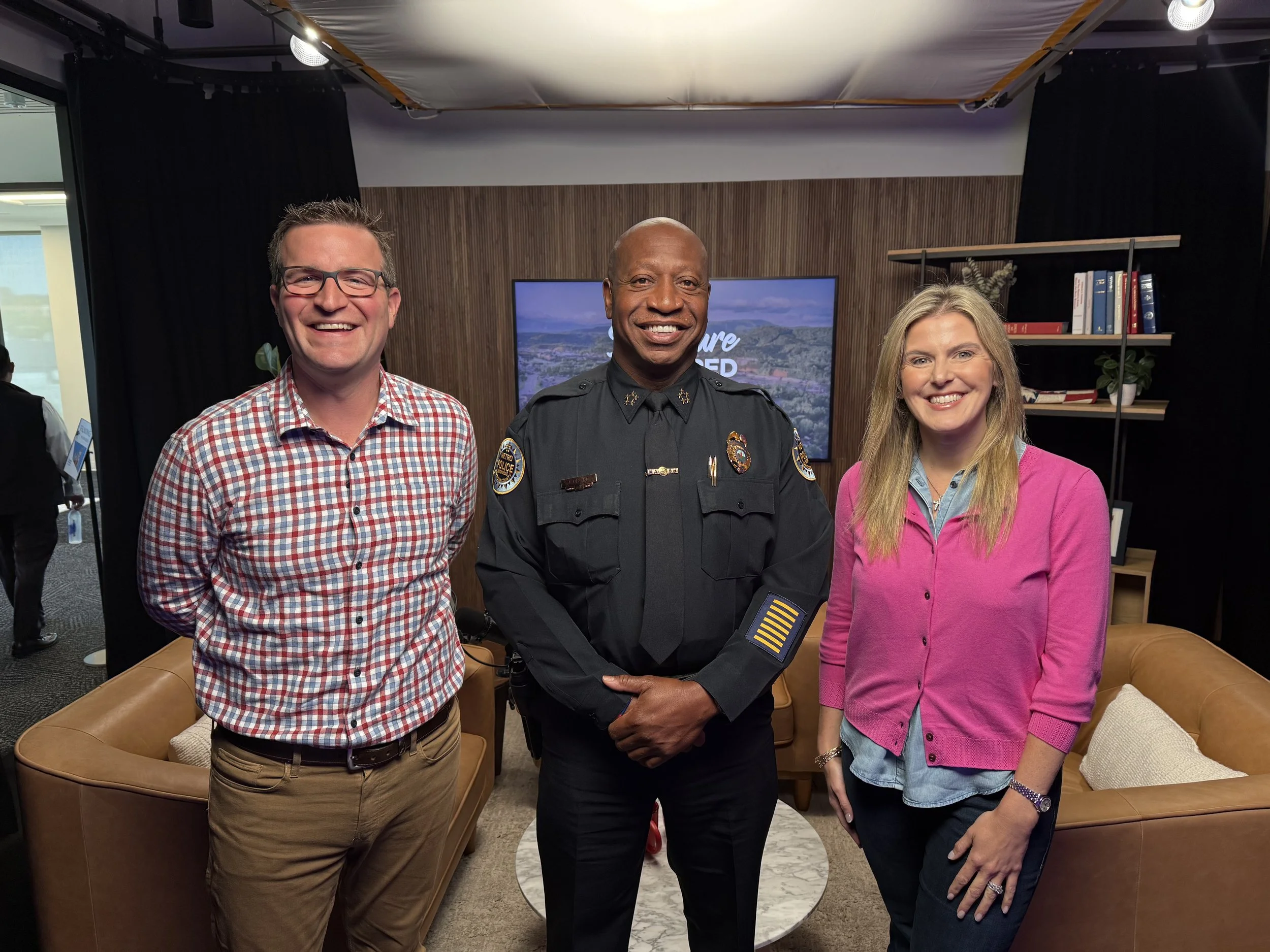Three smiling individuals standing in a room with a television and bookshelves in the background, including a police officer in the center.