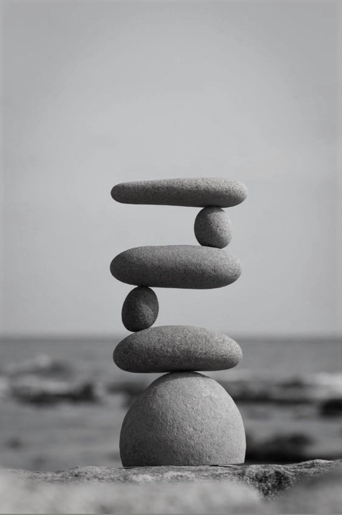 Stack of smooth stones balanced on each other on a beach, with the ocean in the background in black and white.