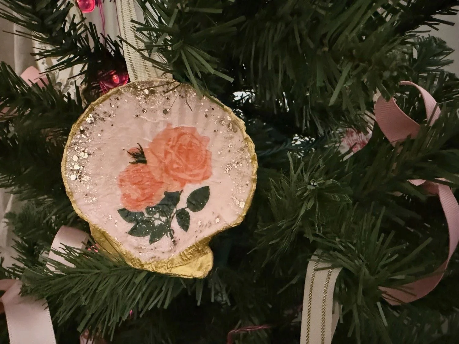 A round ornament with a pink background featuring two orange roses and green leaves, decorated with silver glitter, hanging on a Christmas tree with green branches and pink ribbons.