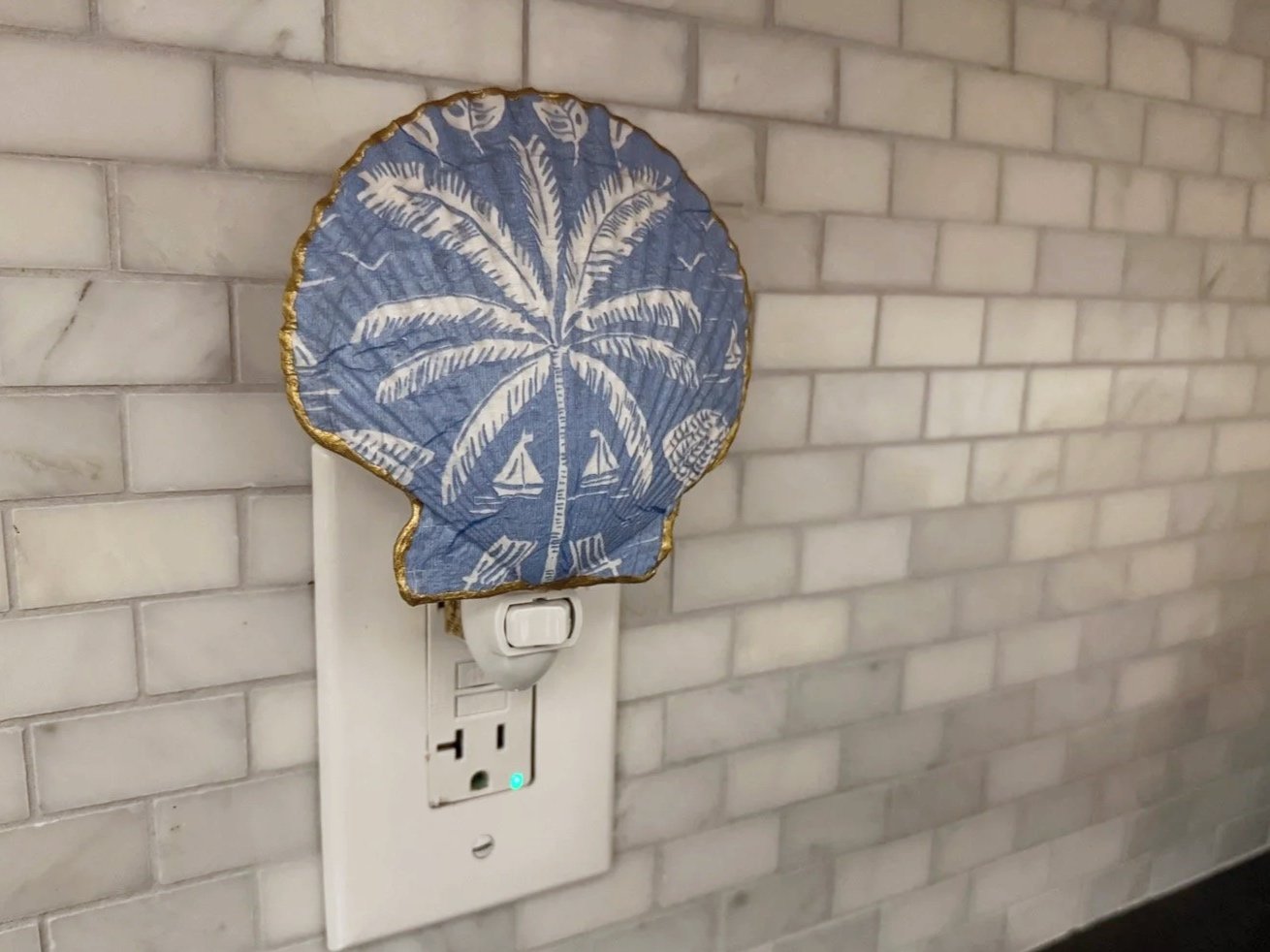A blue and white decorative lampshade hanging on an electrical outlet, with a palm tree and sailboat pattern, attached to a brick wall.