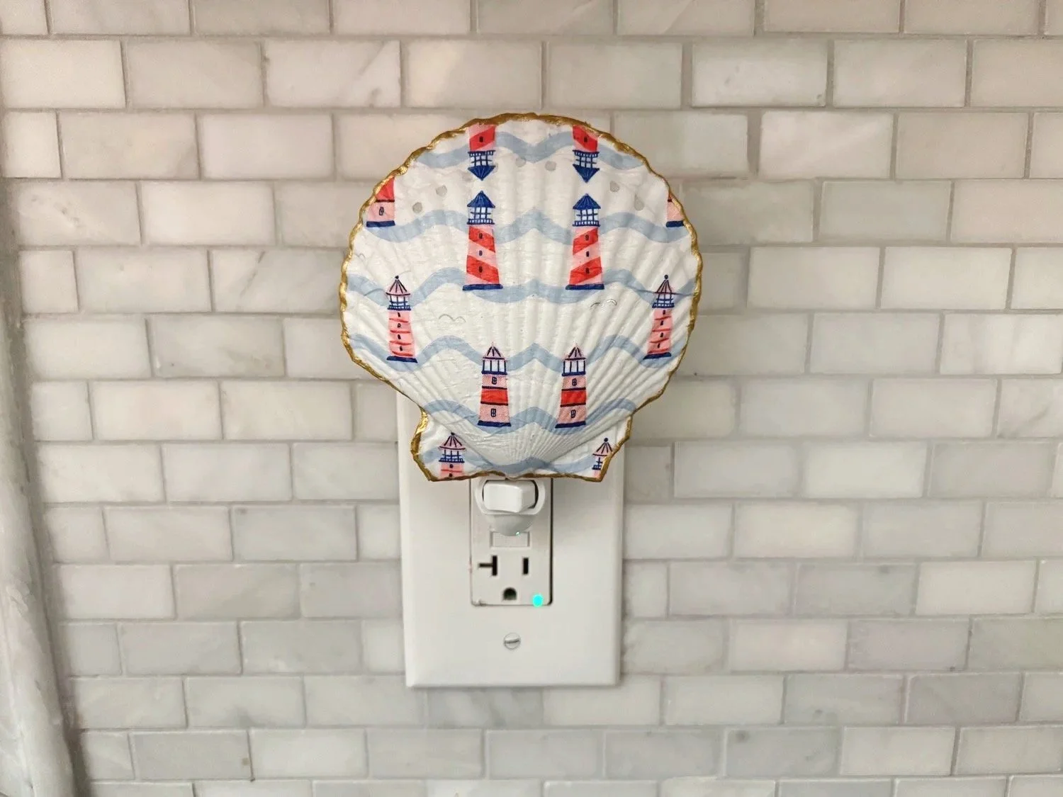 Decorative paper lantern with a lighthouse design hanging over an electrical outlet on a white tiled wall.
