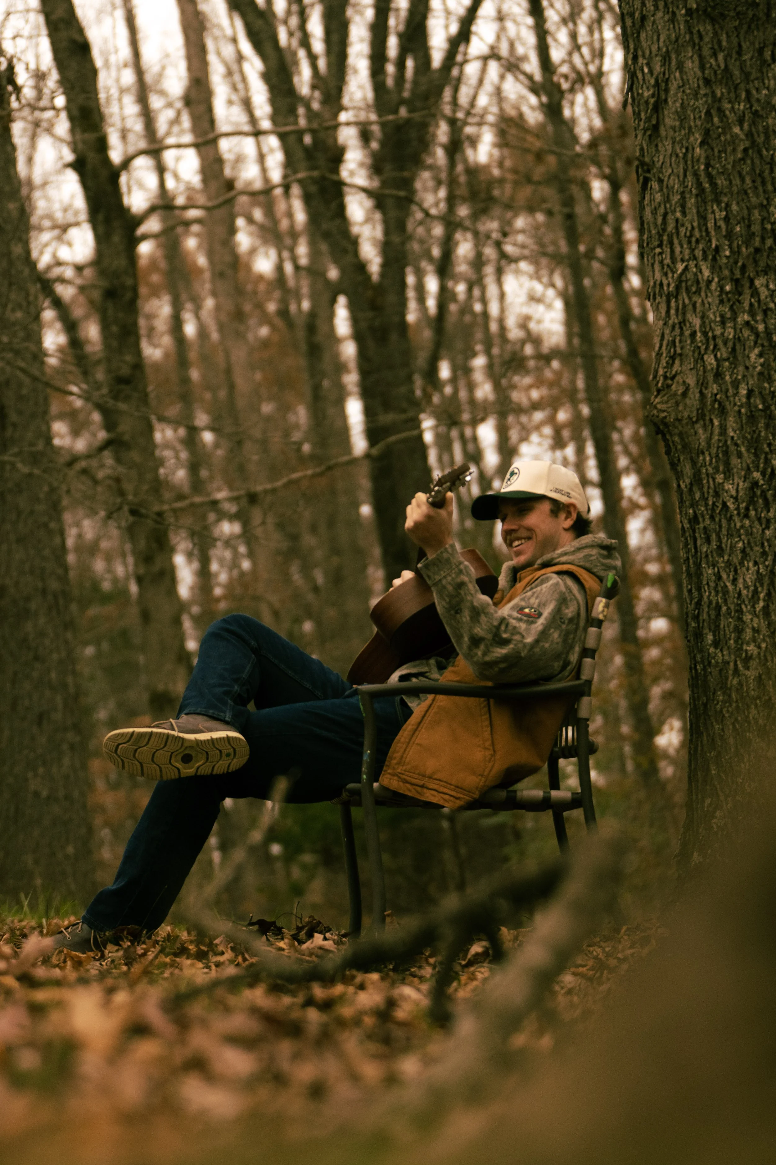 A man sitting on a chair in a forest, playing an acoustic guitar and smiling.