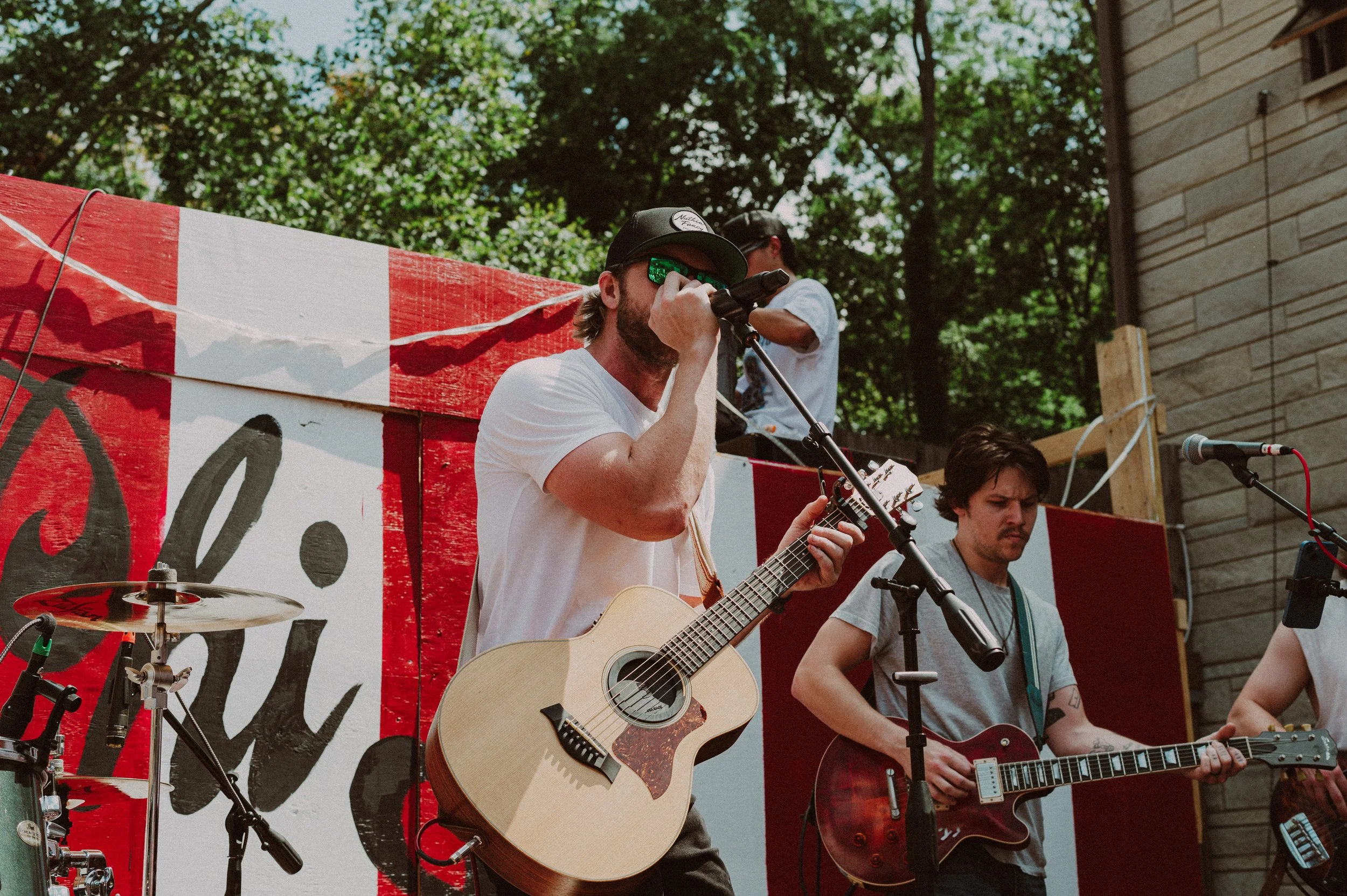 A group of musicians performing outdoors; one playing a guitar and singing into a microphone, another playing an electric guitar, with a drum set partially visible on the left. There is a red and white painted backdrop and greenery in the background.