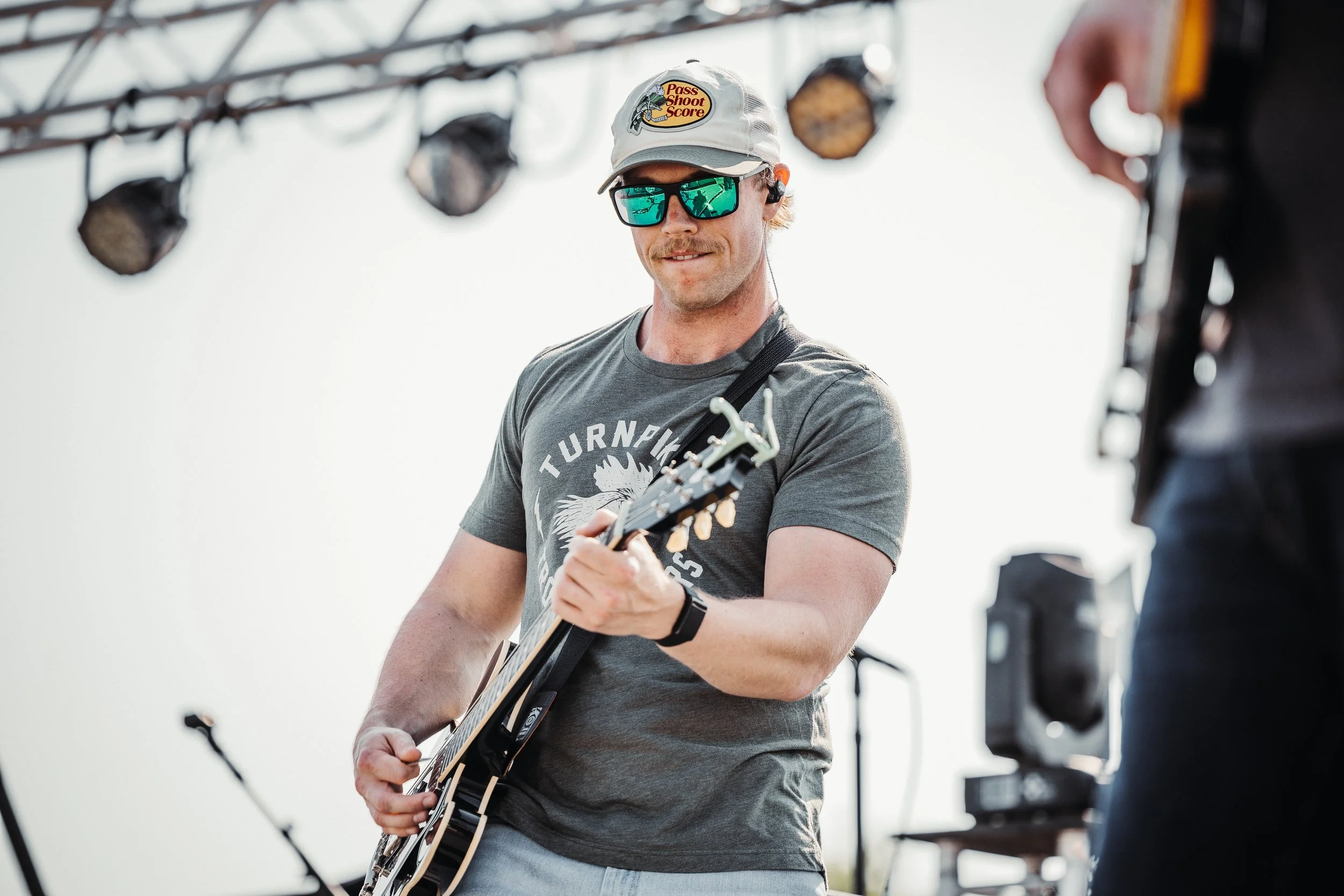 A man wearing a light-colored cap with a logo, sunglasses, and a gray T-shirt playing an electric guitar on stage during a live outdoor performance.