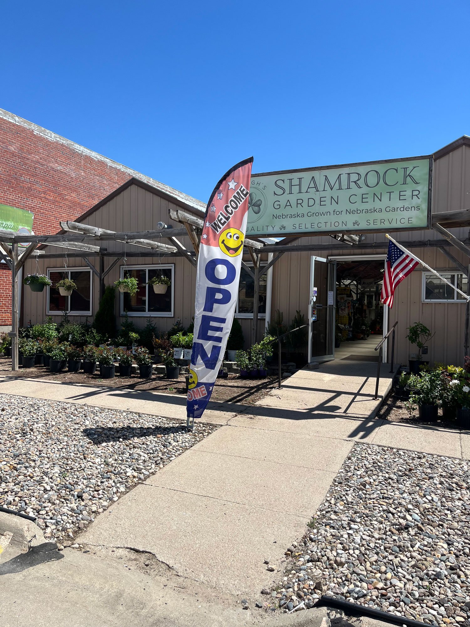 The exterior of a garden center named Shamrock Garden Center with a welcoming flag and American flag near the entrance, surrounded by plants and gravel, under a clear blue sky.
