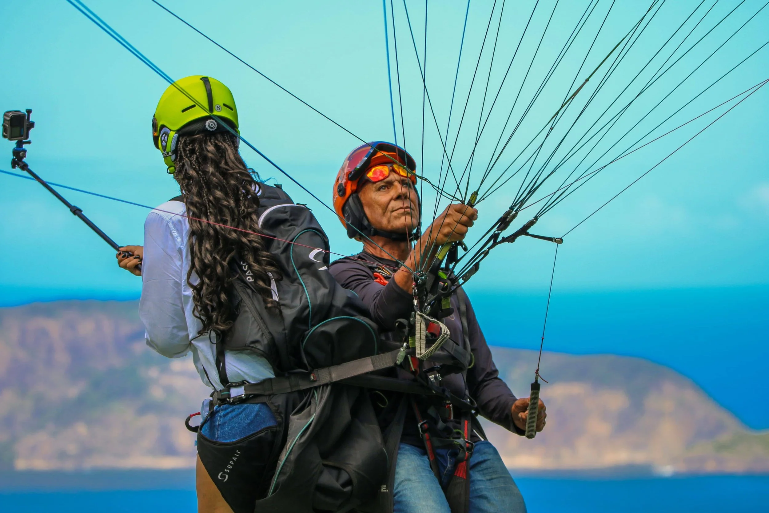 Homem com capacete laranja controlando parapente em céu azul, ao lado de uma mulher com capacete verde, ambos equipados com mochilas de voos e roupas adequadas para parapente, ao fundo paisagem de água e colinas.