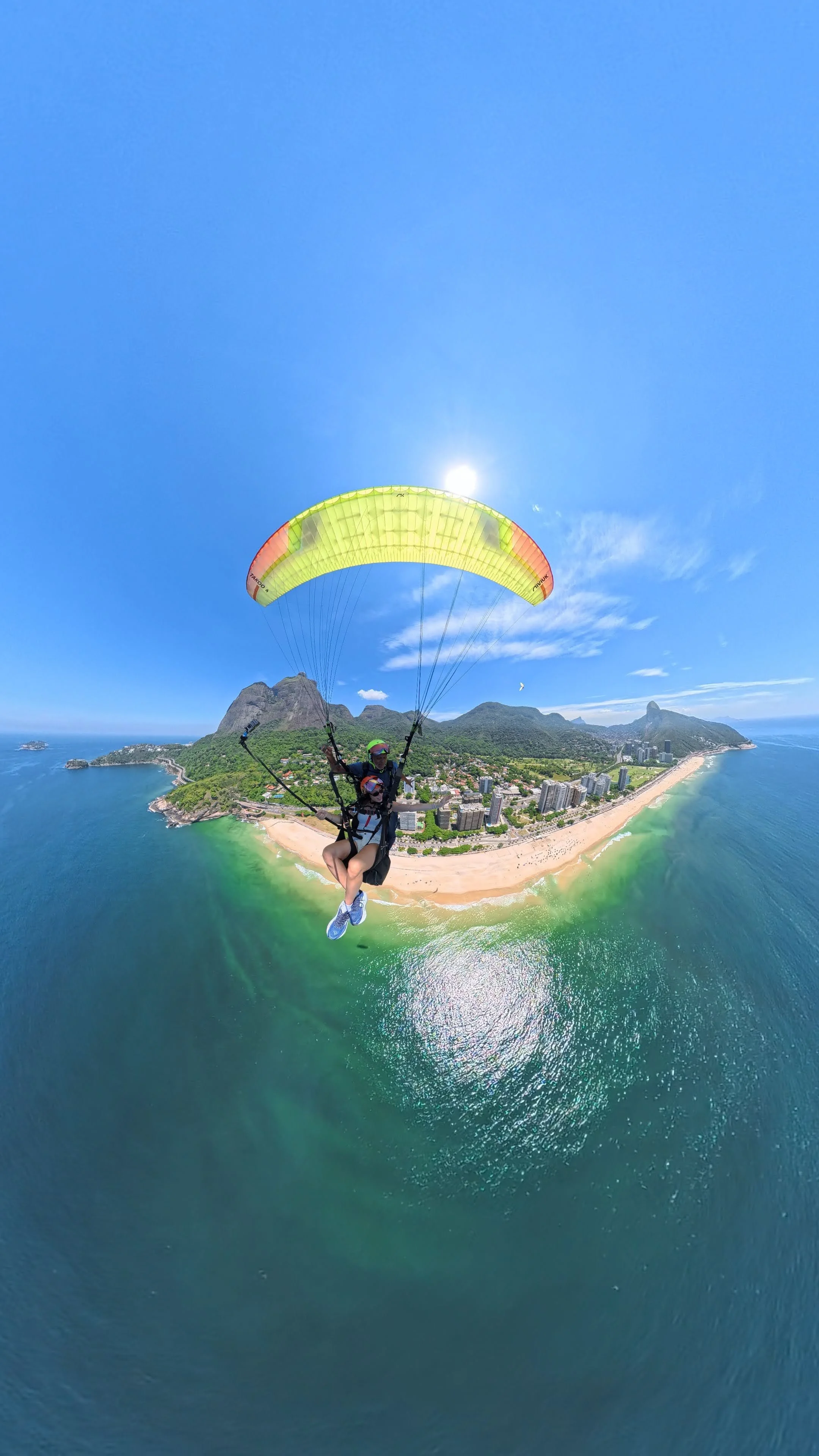Vôo de paramotor sobre o litoral com praia, prédios e montanhas ao fundo, céu azul com sol brilhando.