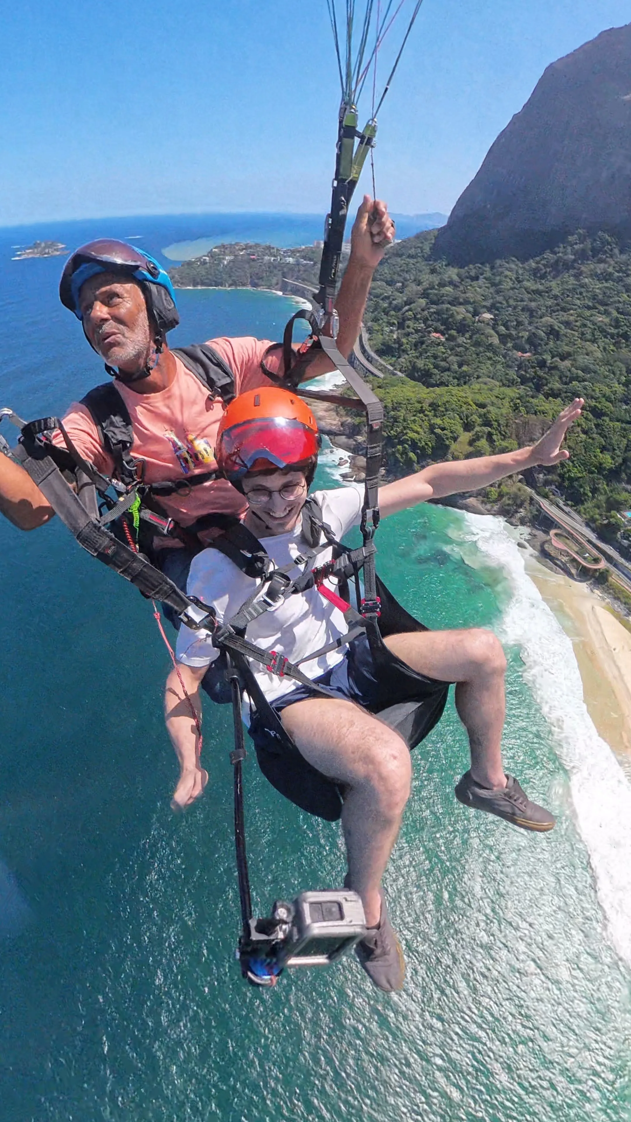 Dois homens fazendo parapente sobre a praia com vista para o mar e as montanhas.