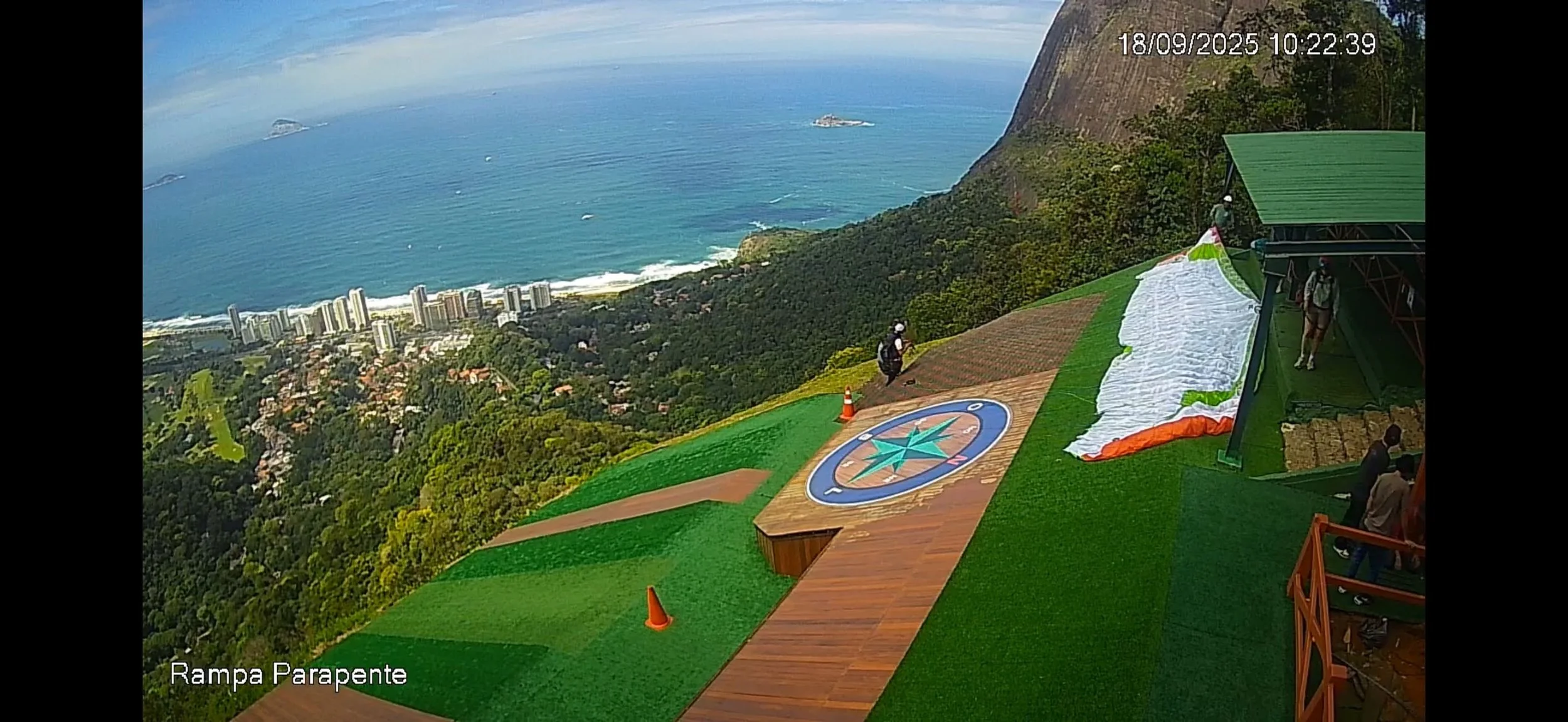 Vista do topo de uma montanha com uma rampa de parapente, áreas verdes, uma área de madeira, cones de trânsito, uma lona branca desdobrada e uma estrutura de sustentação. Ao fundo, a cidade costeira com prédios altos e o mar com pequenas ilhas, ao la