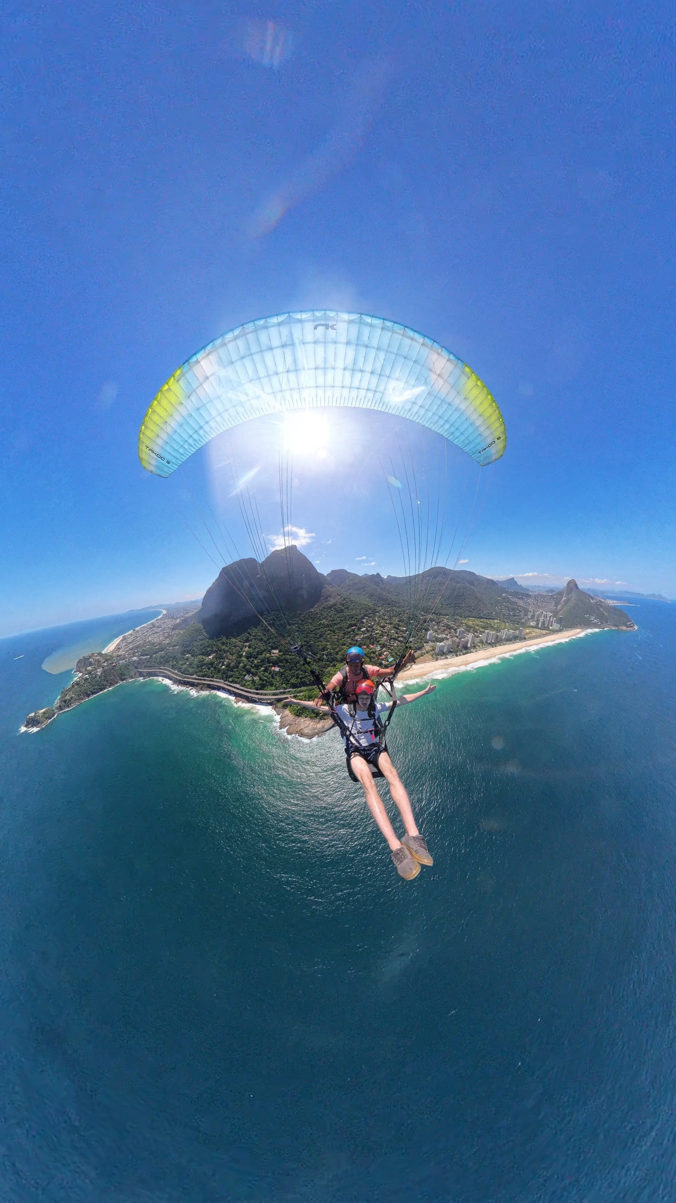 Dupla em parapente sobre a costa de uma cidade litorânea com praias, montanhas e céu azul.
