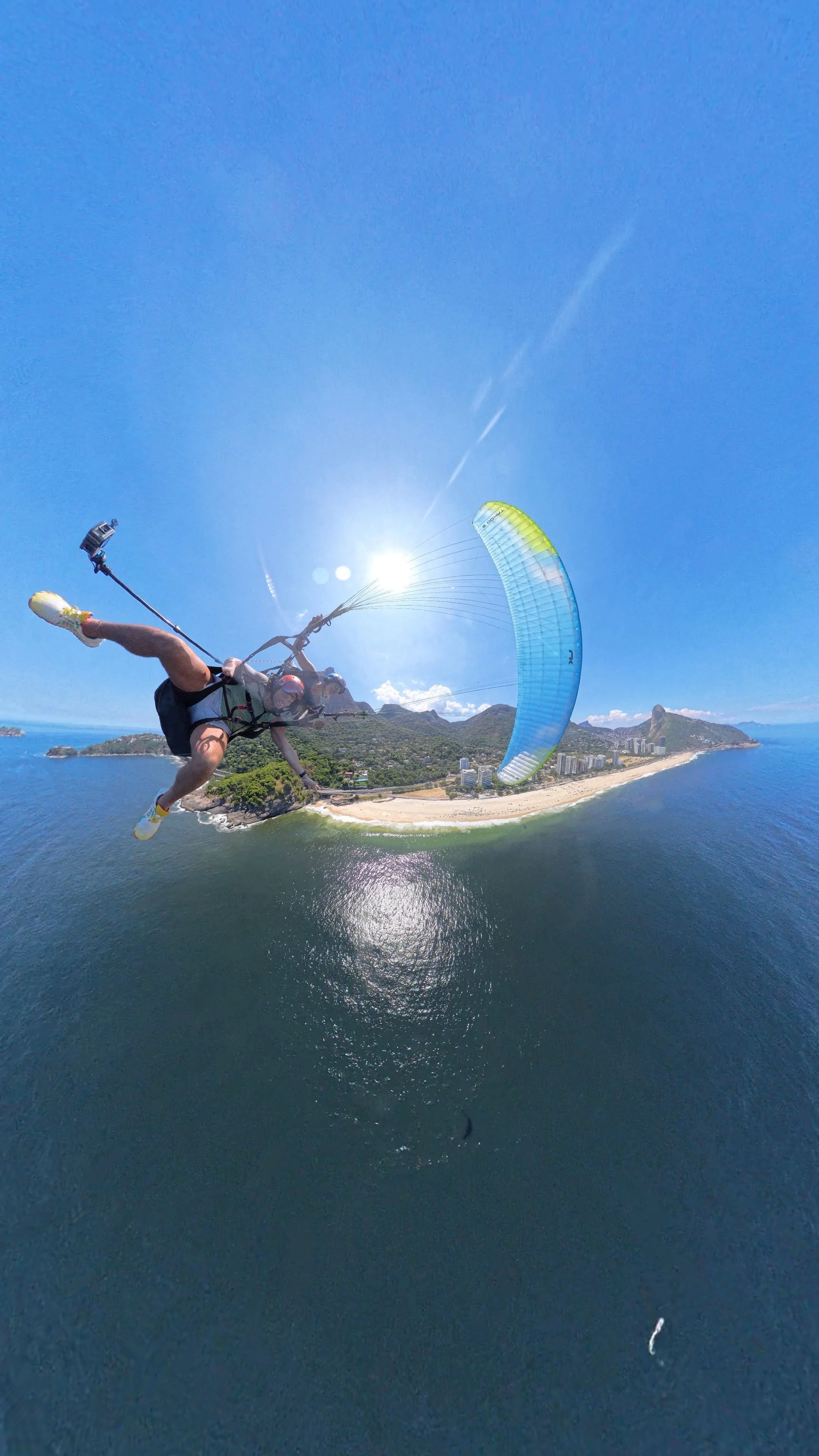 Pessoa realizando parapente sobre uma praia com mar azul e uma linha do horizonte ao fundo em uma tarde ensolarada.