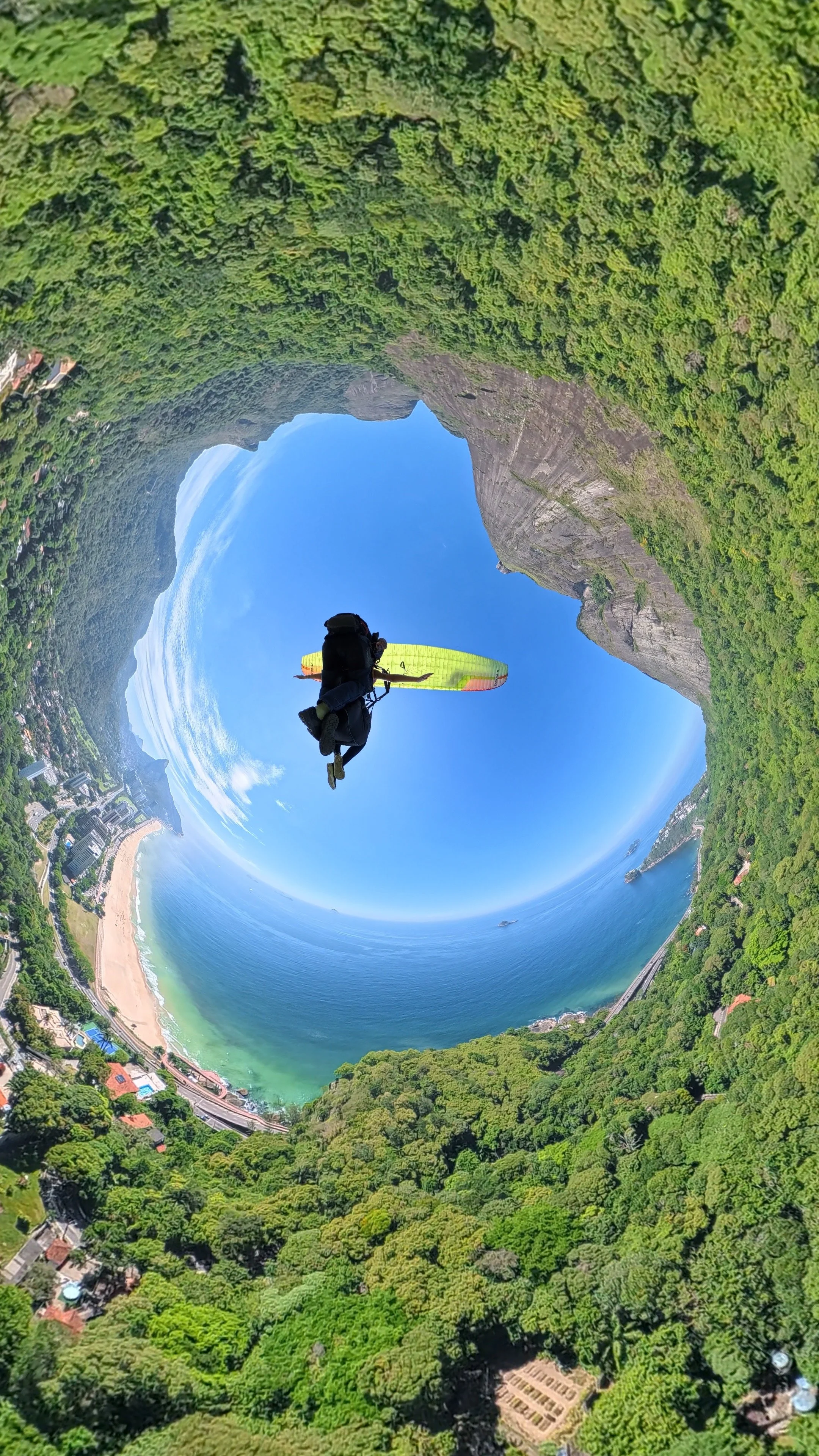Aprendiz de parapente voando sobre uma paisagem costeira com praias, cidade e árvores verdes, capturado com efeito de mundo em miniatura.