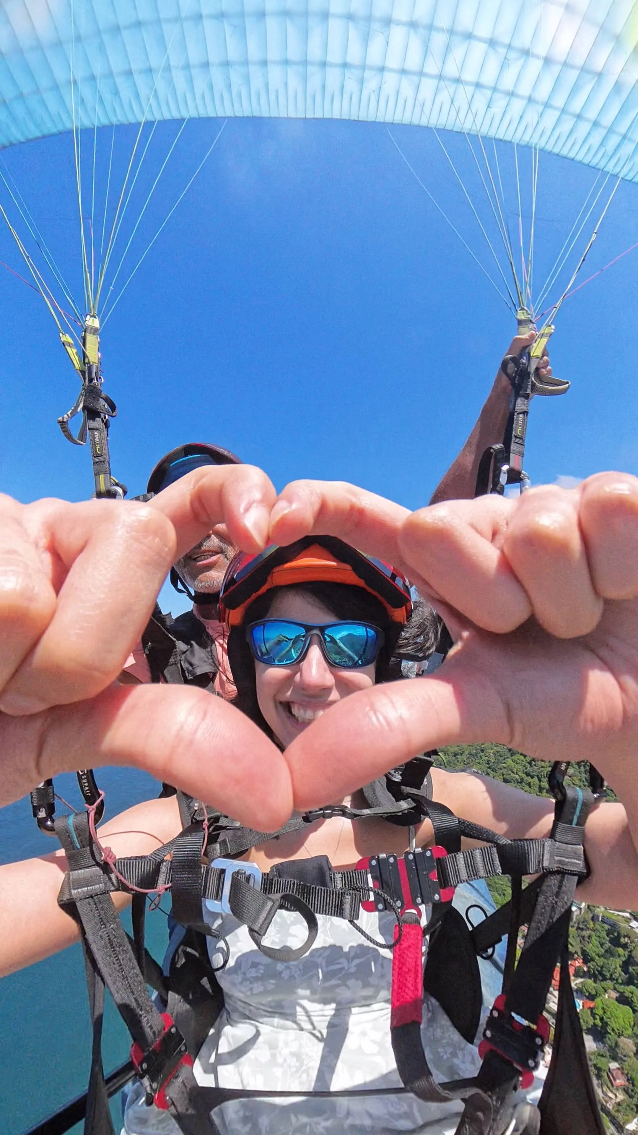 Pessoa fazendo parapente, formando um coração com as mãos, vestida com equipamento de segurança e óculos de sol, com céu azul ao fundo.