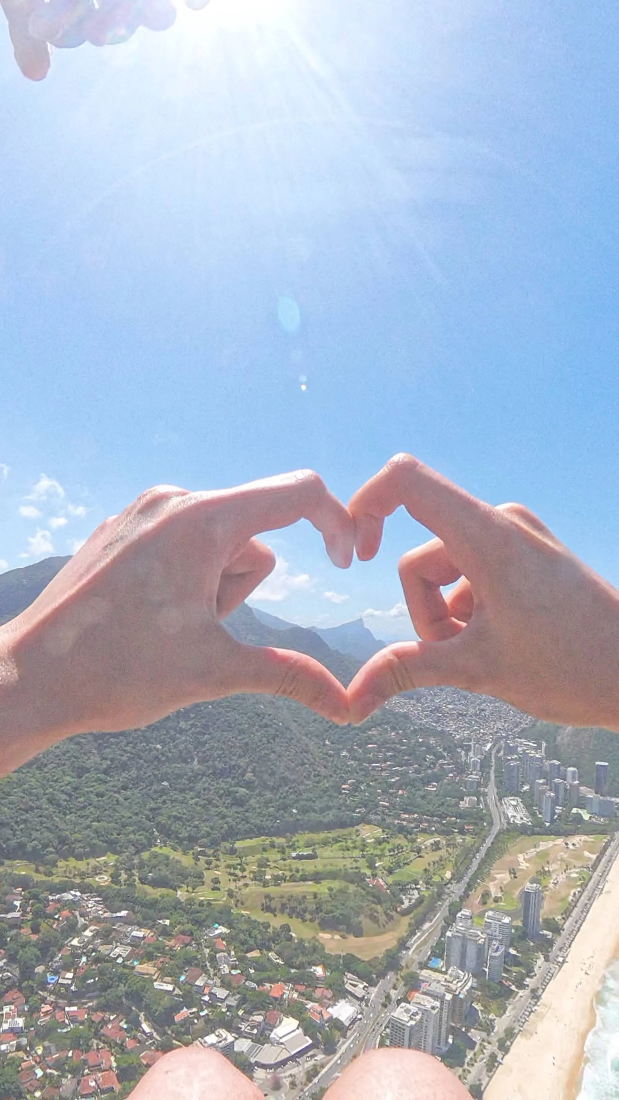 Pessoa formando um coração com as mãos durante um dia ensolarado, com vista para uma cidade costeira e montanhas ao fundo.