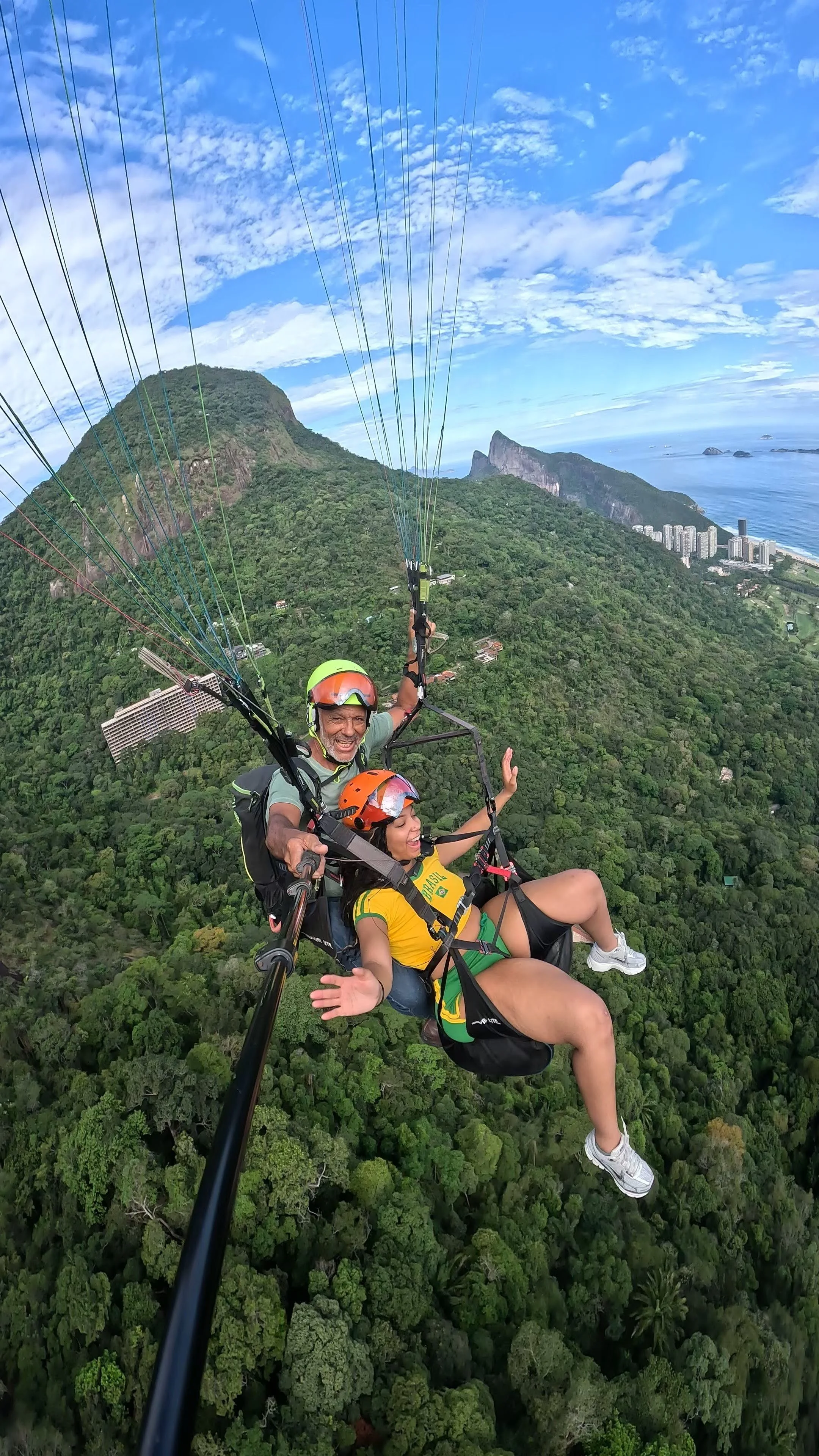 Duas pessoas fazendo parapente sobre uma floresta, com montanhas ao fundo e céu azul com nuvens.