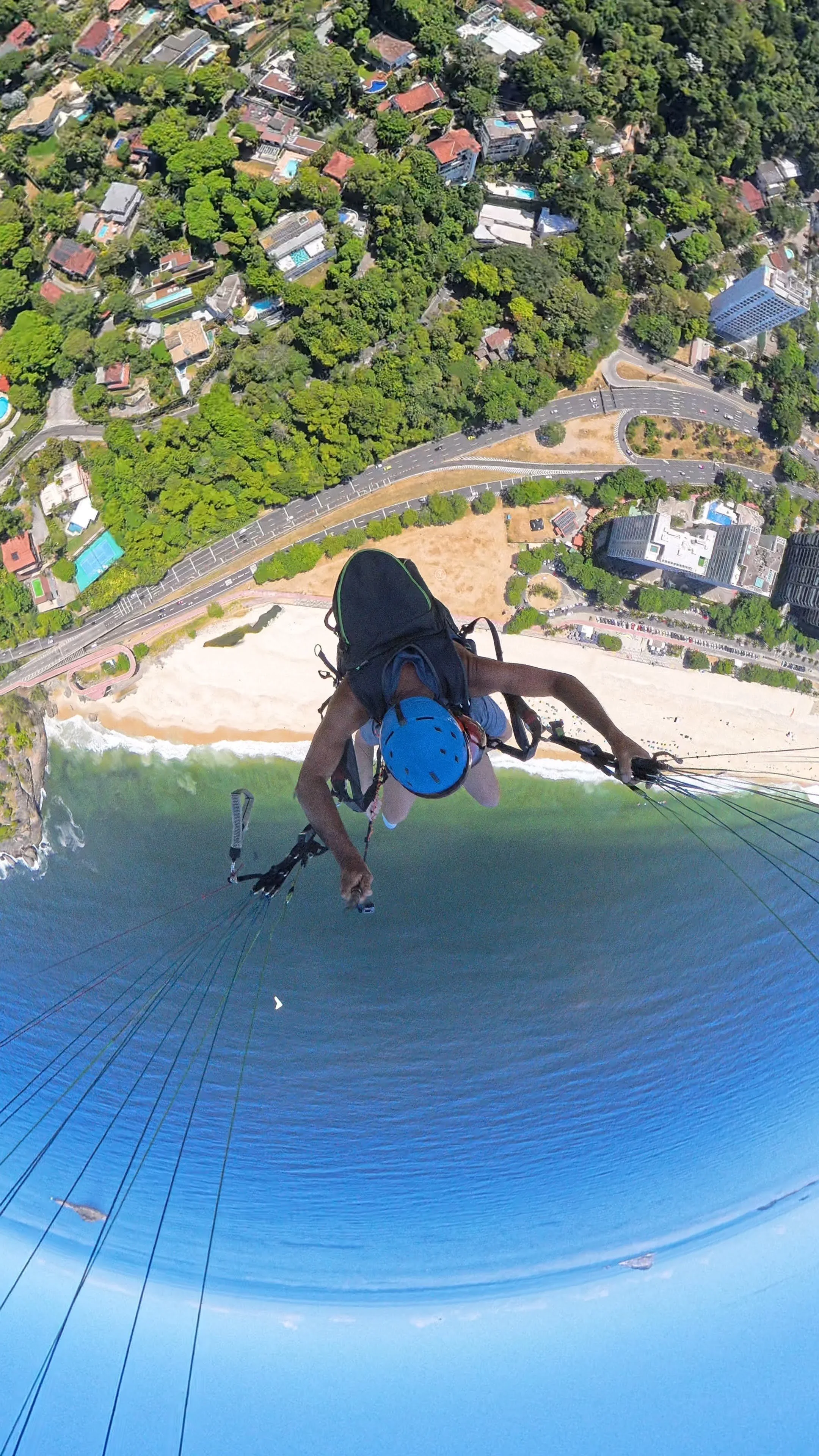 Pessoa praticando voo de parapente acima de uma praia com água azul, areia e áreas verdes, com uma cidade ao fundo.