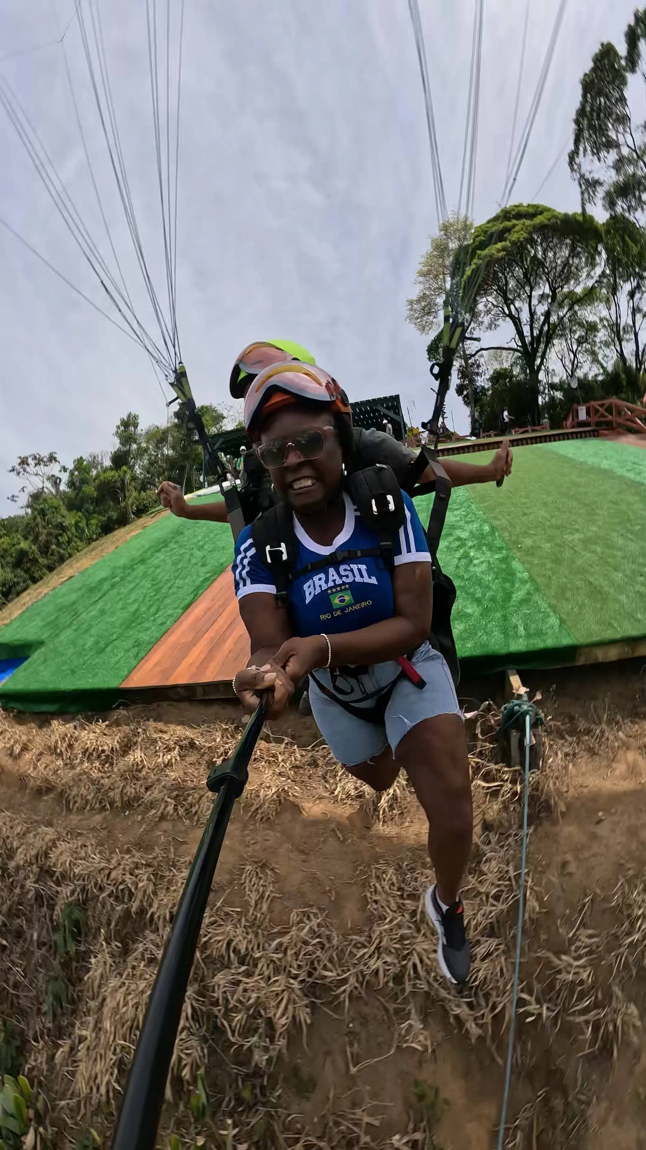Pessoa fazendo selfie com ação de paraquedismo, usando capacete, óculos de proteção, camiseta do Brasil e mochila, ao sair do avião em voo de paraquedismo.