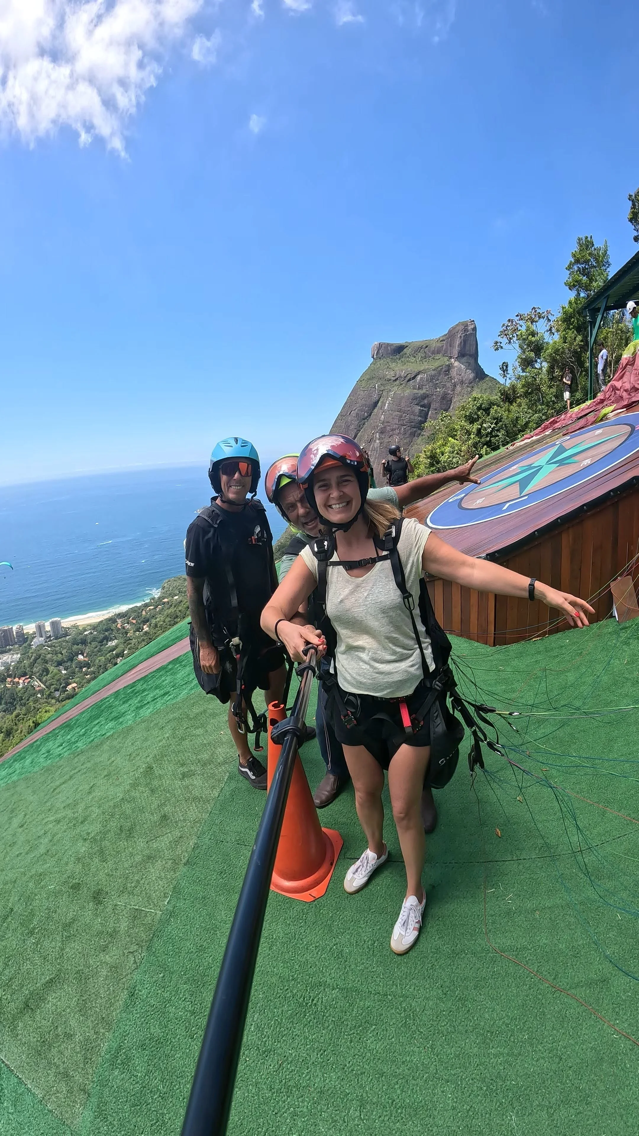 Grupo de pessoas com equipamentos de paraquedismo sorrindo para a câmera na encosta de uma montanha, com o Cristo Redentor ao fundo, em uma bela paisagem com céu azul e vista para o mar.