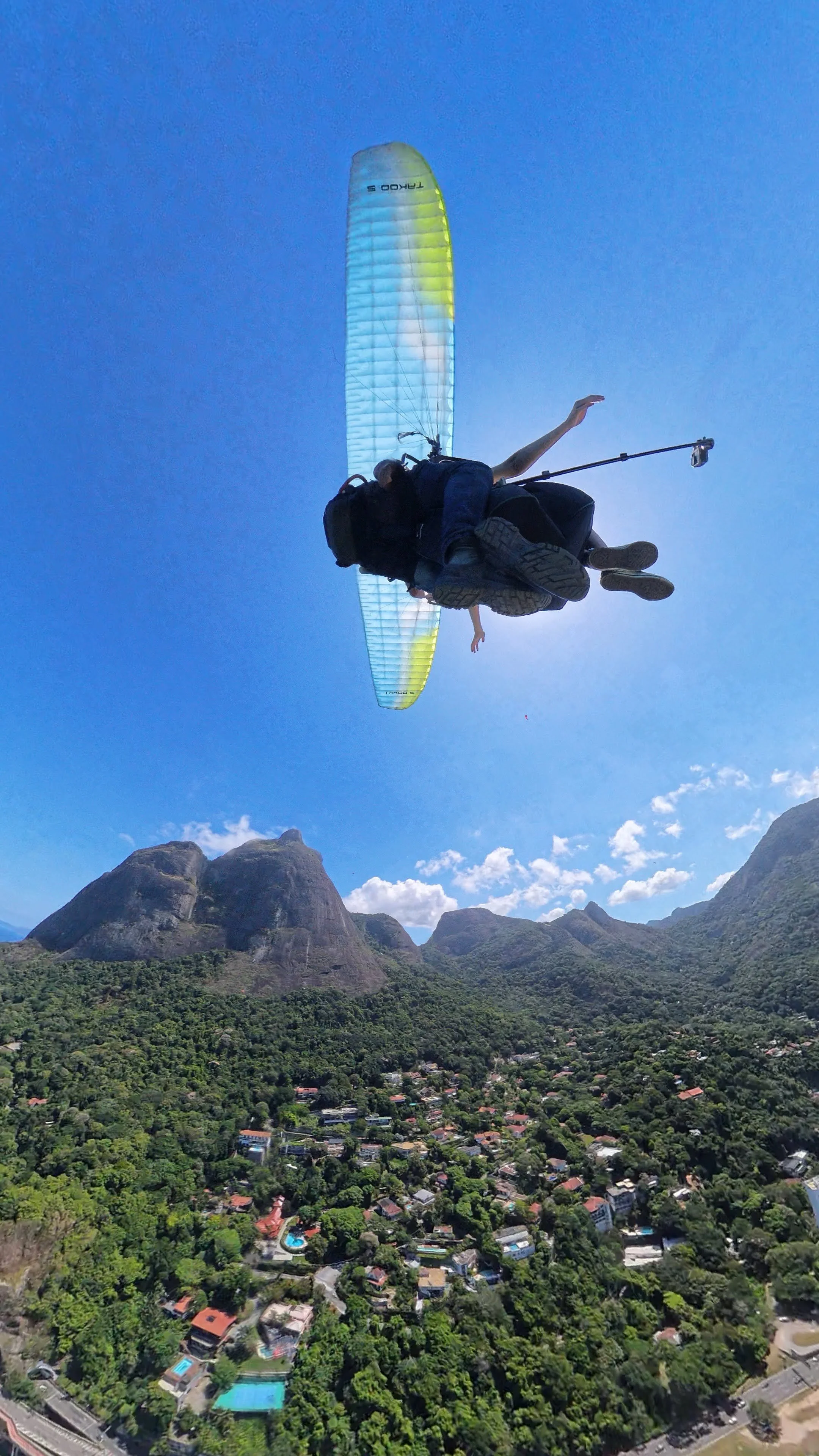 Paraglider voando sobre uma paisagem de montanhas verdes e uma cidade com casas e piscinas ao sol.
