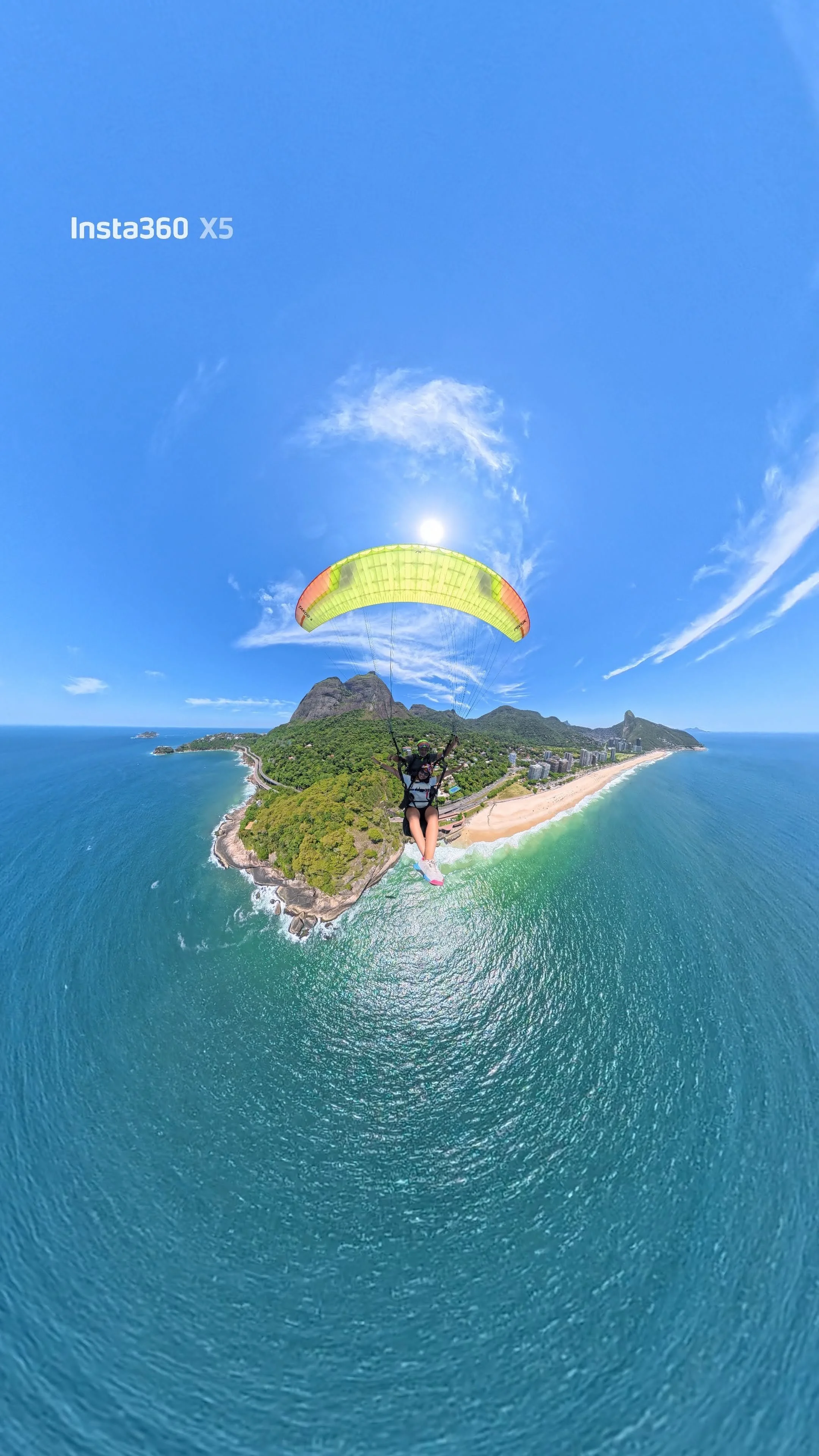 Pessoa praticando parapente acima de uma costa com mar, praia e montanhas ao fundo durante o dia.