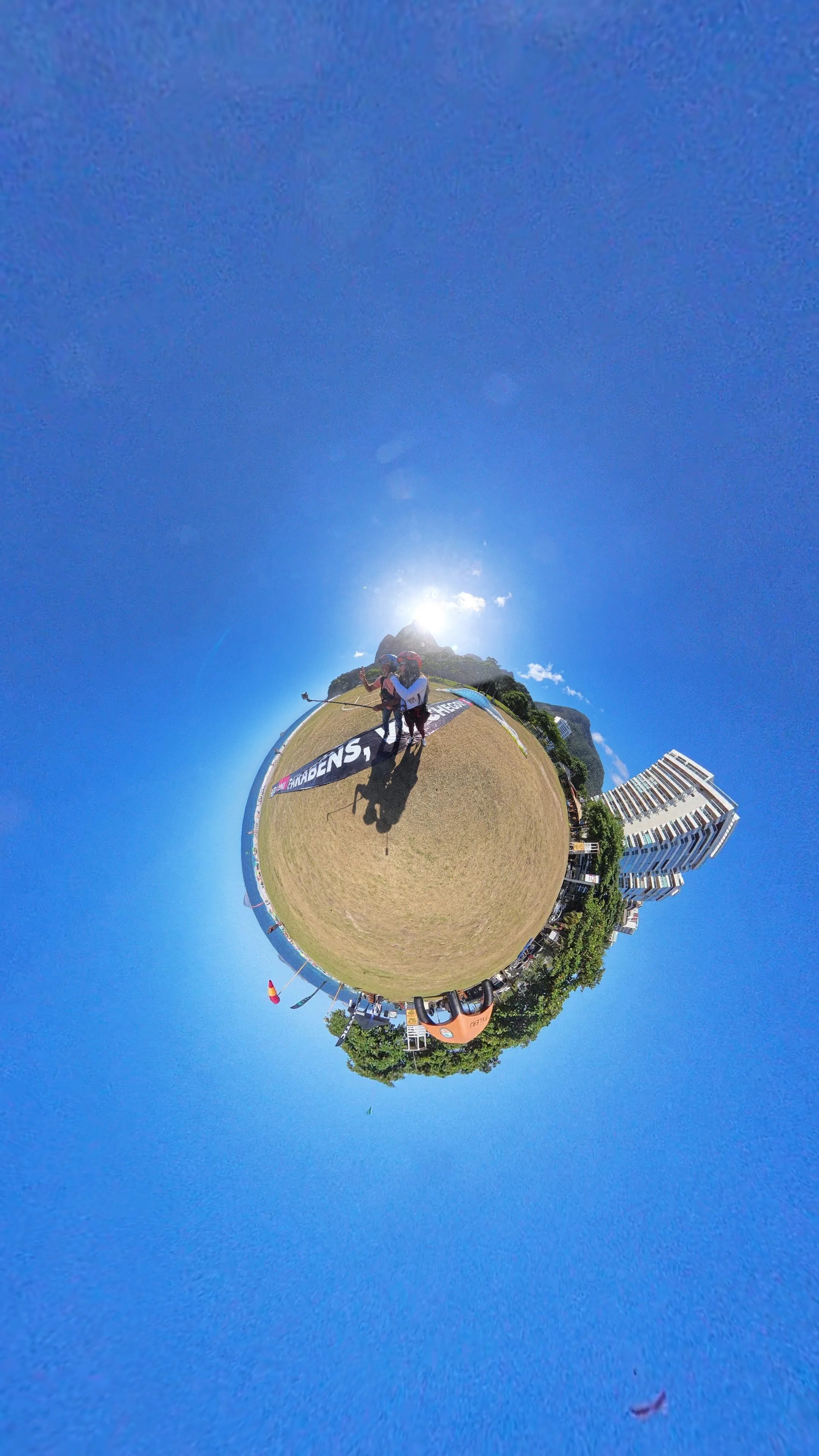 Imagem em estilo de planeta miniatura, mostrando uma equipe de pessoas ao ar livre, com edifícios e árvores ao fundo, uma bandeira e um céu azul sob o sol.