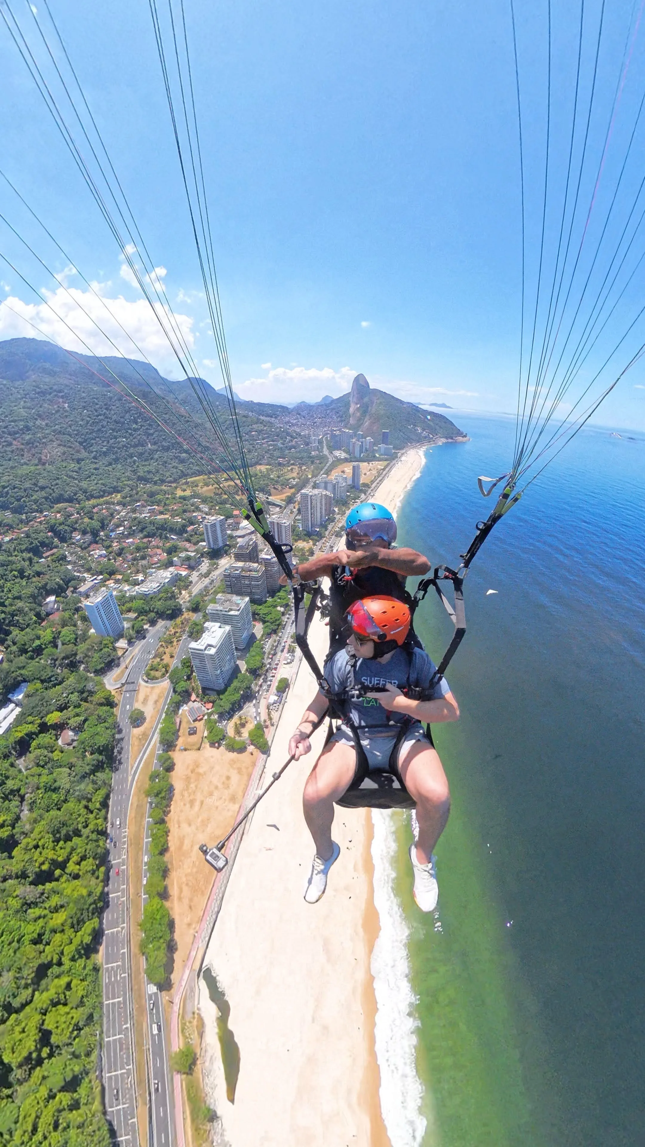 Pessoas fazendo parapente sobre uma praia com vista para a cidade e o mar ao fundo, com céu azul e algumas nuvens.