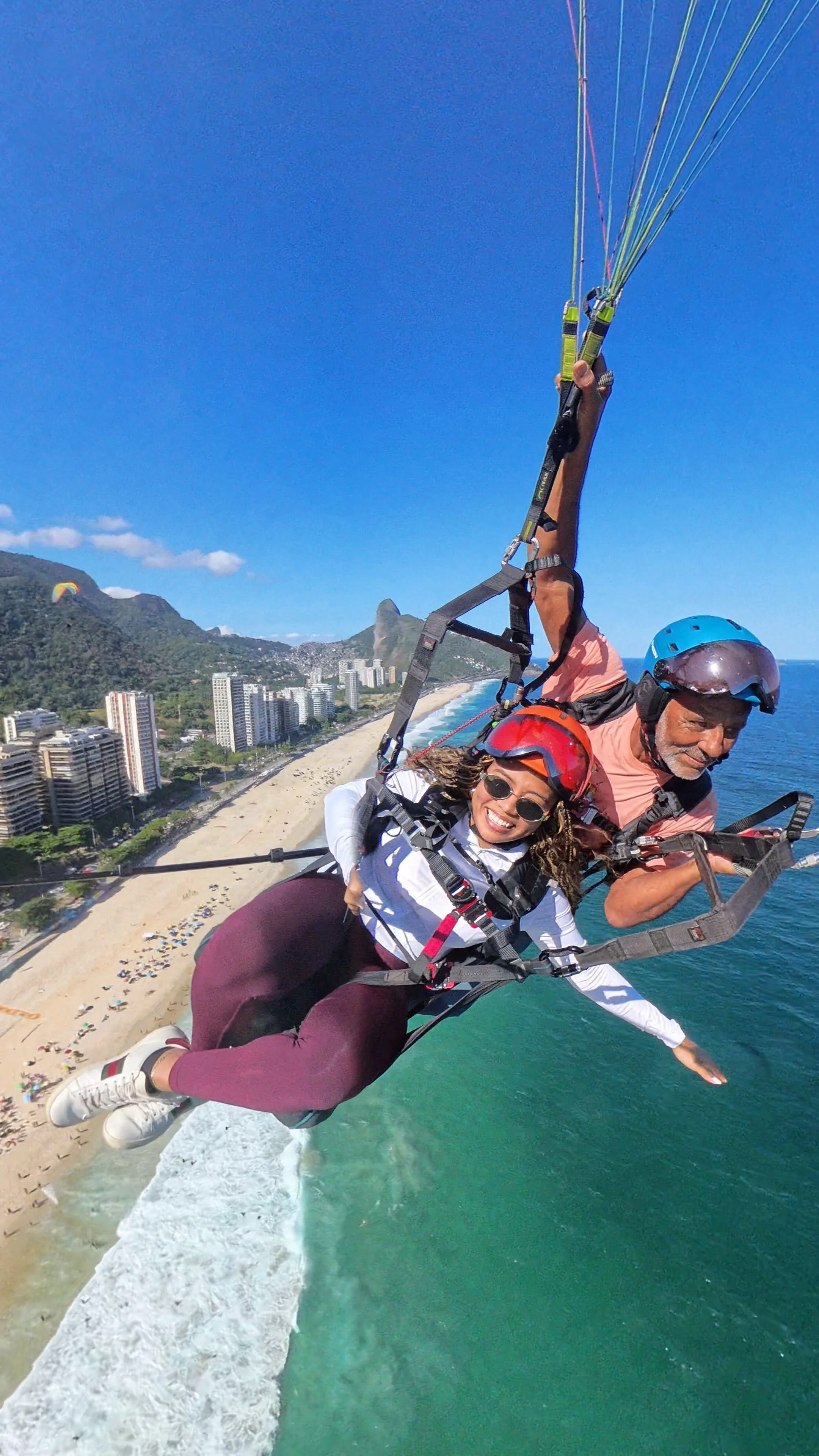 Casal fazendo parapente sobre a praia, com edifícios e montanhas ao fundo.