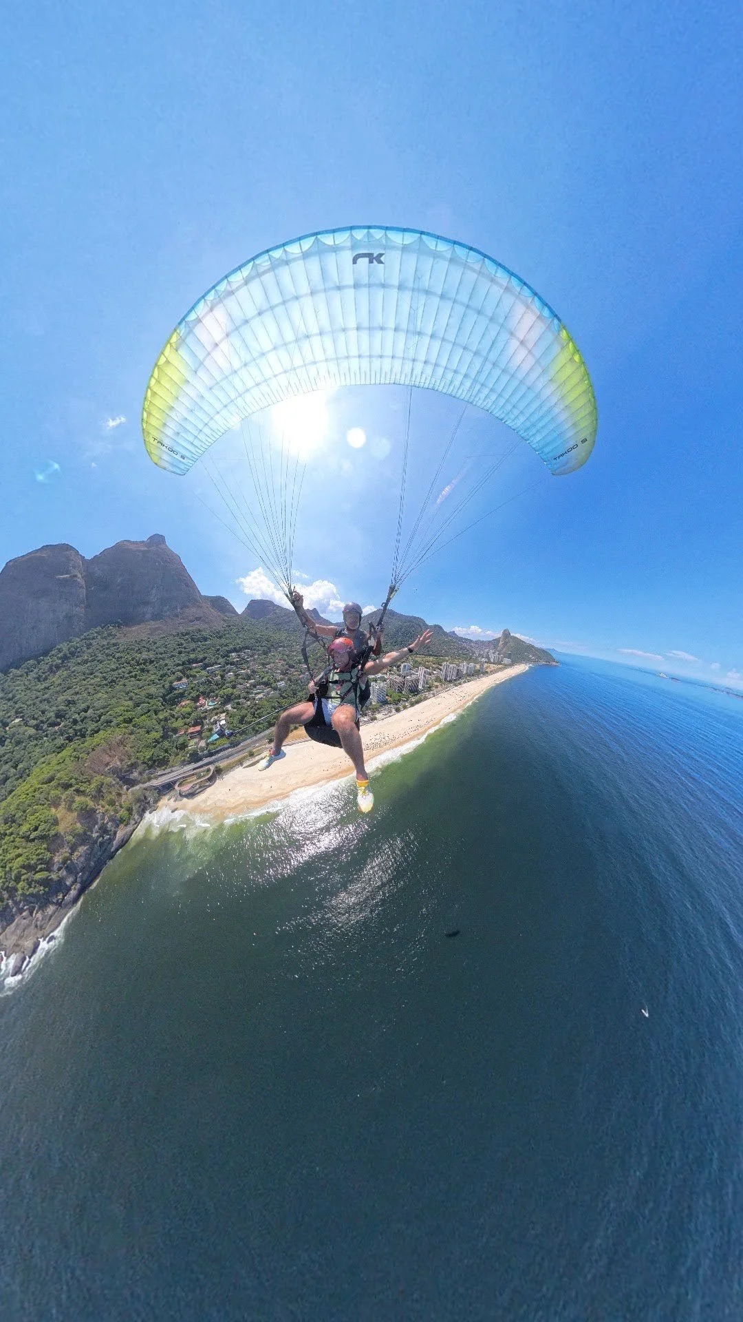 Duas pessoas fazendo parapente sobre praia com montanhas ao fundo, céu claro e sol brilhando.