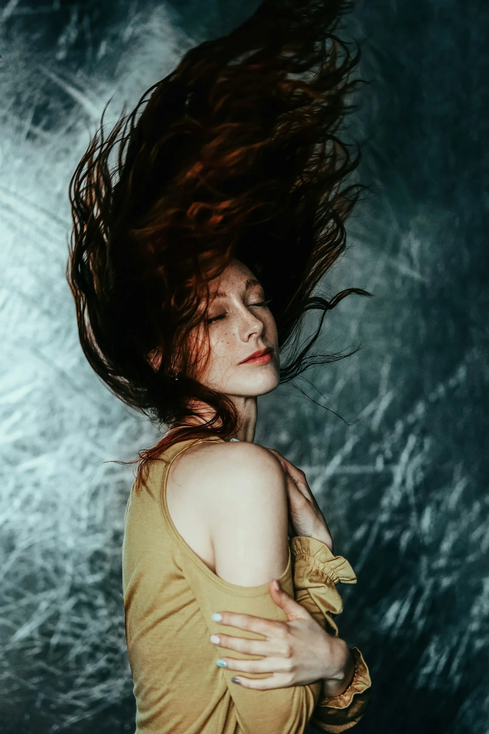 Woman with one arm wrapped around herself, one hand on her chest, her eyes closes and hair blowing in the wind