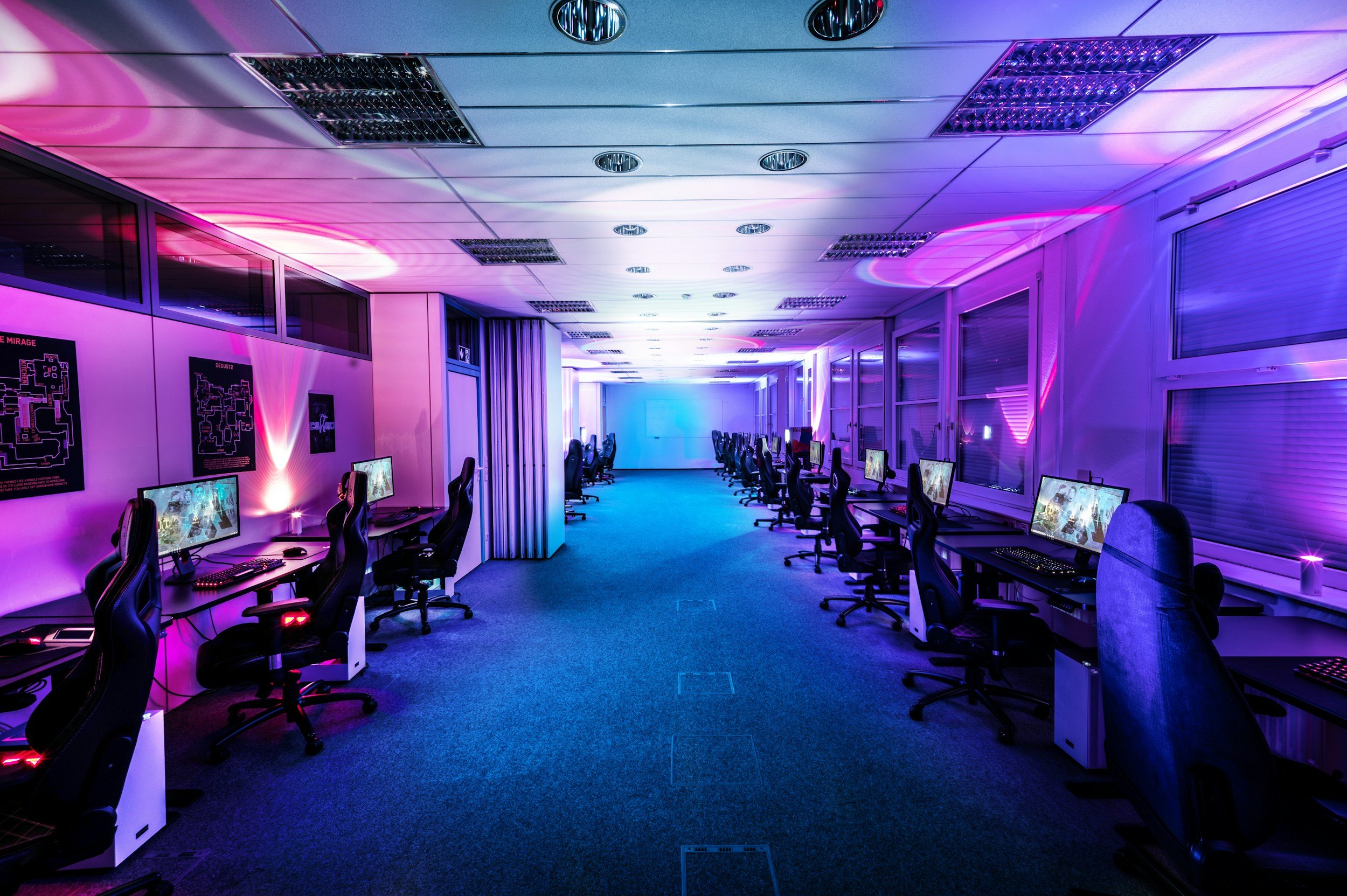 An indoor gaming or computer lab room with multiple computer stations lined up along both sides. The room is illuminated with vibrant pink and purple LED lighting on the ceiling and walls, creating a futuristic ambiance.