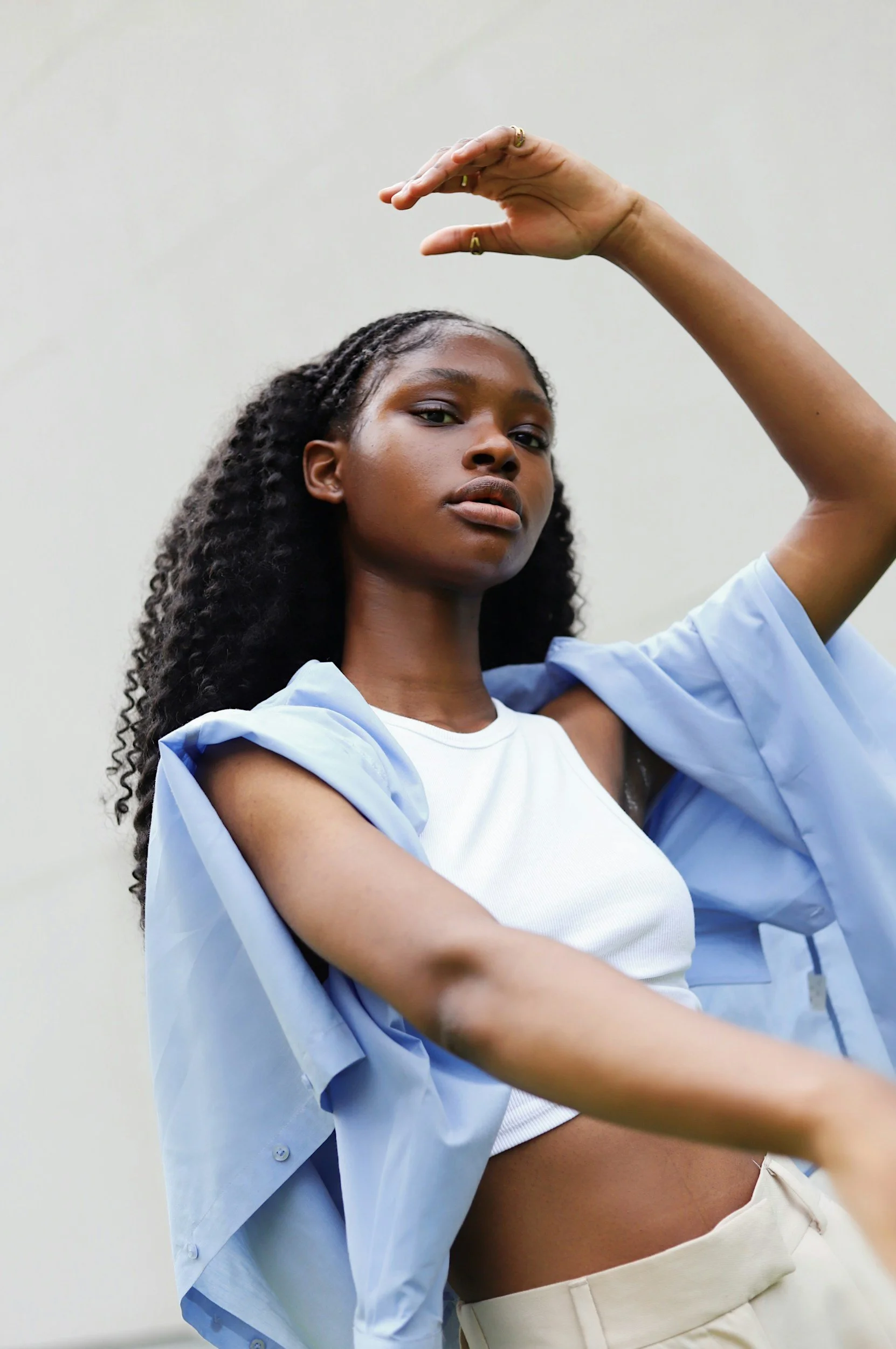 A woman with dark skin and curly hair is posing with one arm raised above her head, looking directly at the camera, wearing a light blue shirt over a white top against a plain wall.