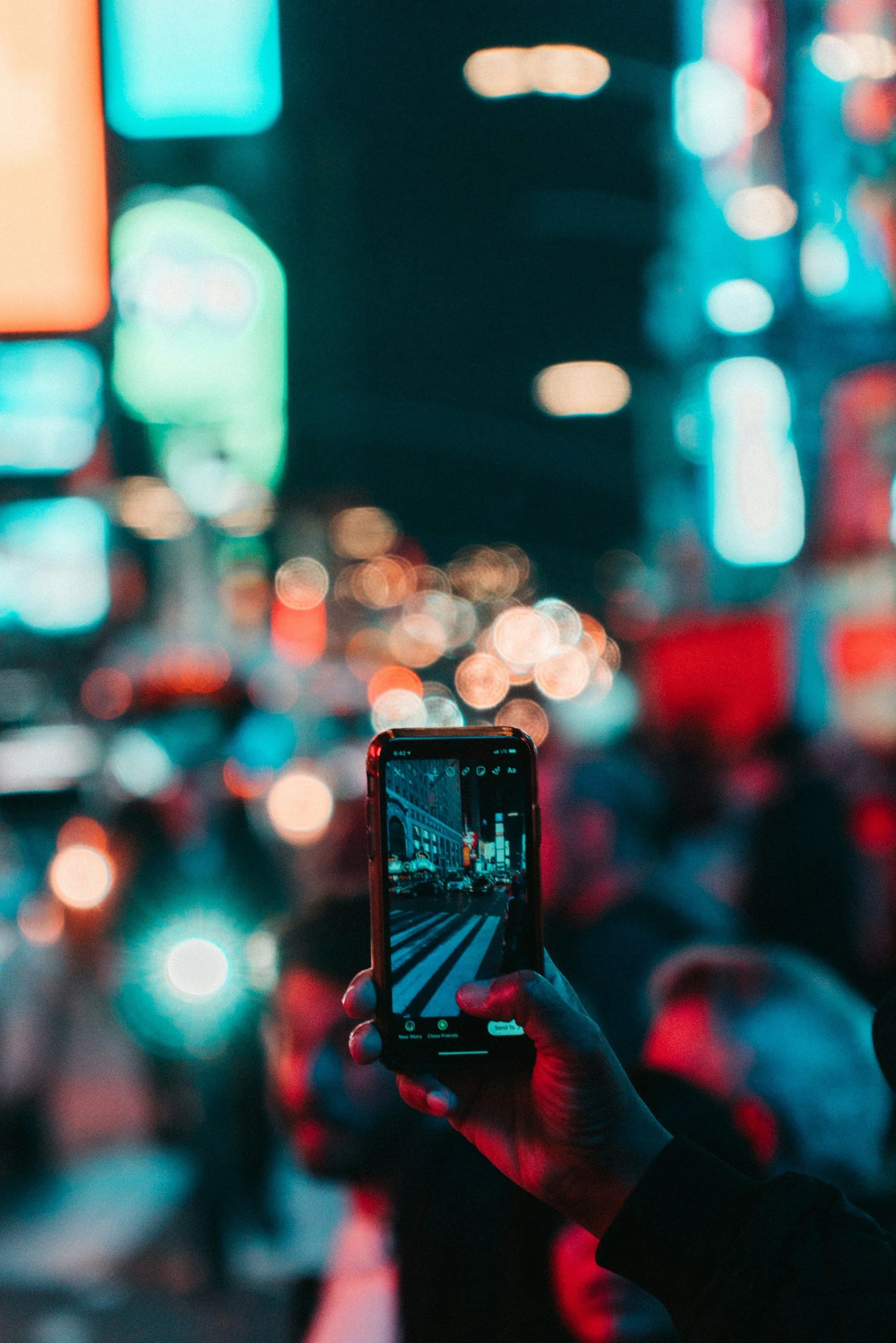 Person holding a smartphone taking a picture of a city street at night, with brightly lit buildings and blurred colorful lights.