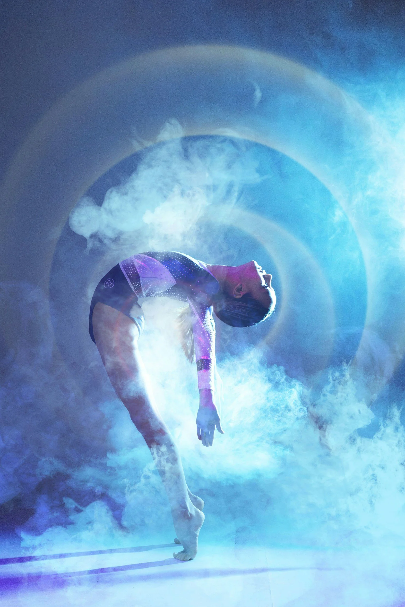 A person performing a handstand with one leg bent and the other extended, surrounded by blue smoke and a circular rainbow-like light in a dark setting.