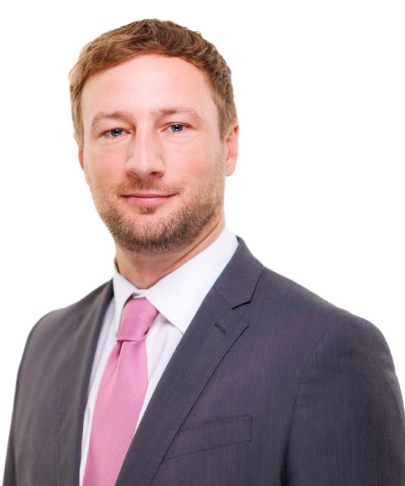 A man wearing a dark suit, white shirt, and pink tie, looking at the camera with a slight smile, against a plain white background.