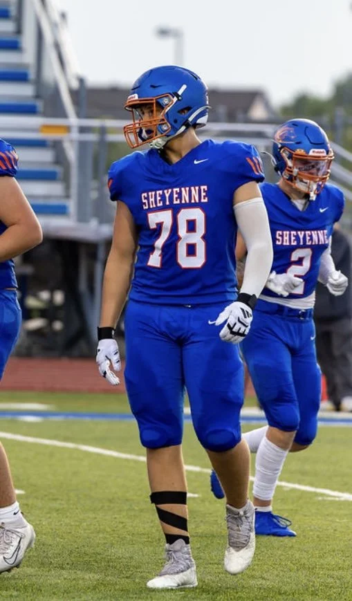 Two female football players in blue uniforms with 'SHEYENNE' written on the jerseys, wearing helmets, standing on a football field during daytime.