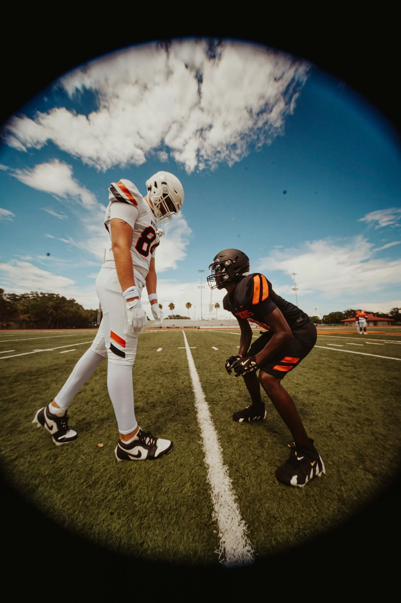Two football players face each other on a field, one in a white uniform and the other in a black uniform, under a partly cloudy sky.
