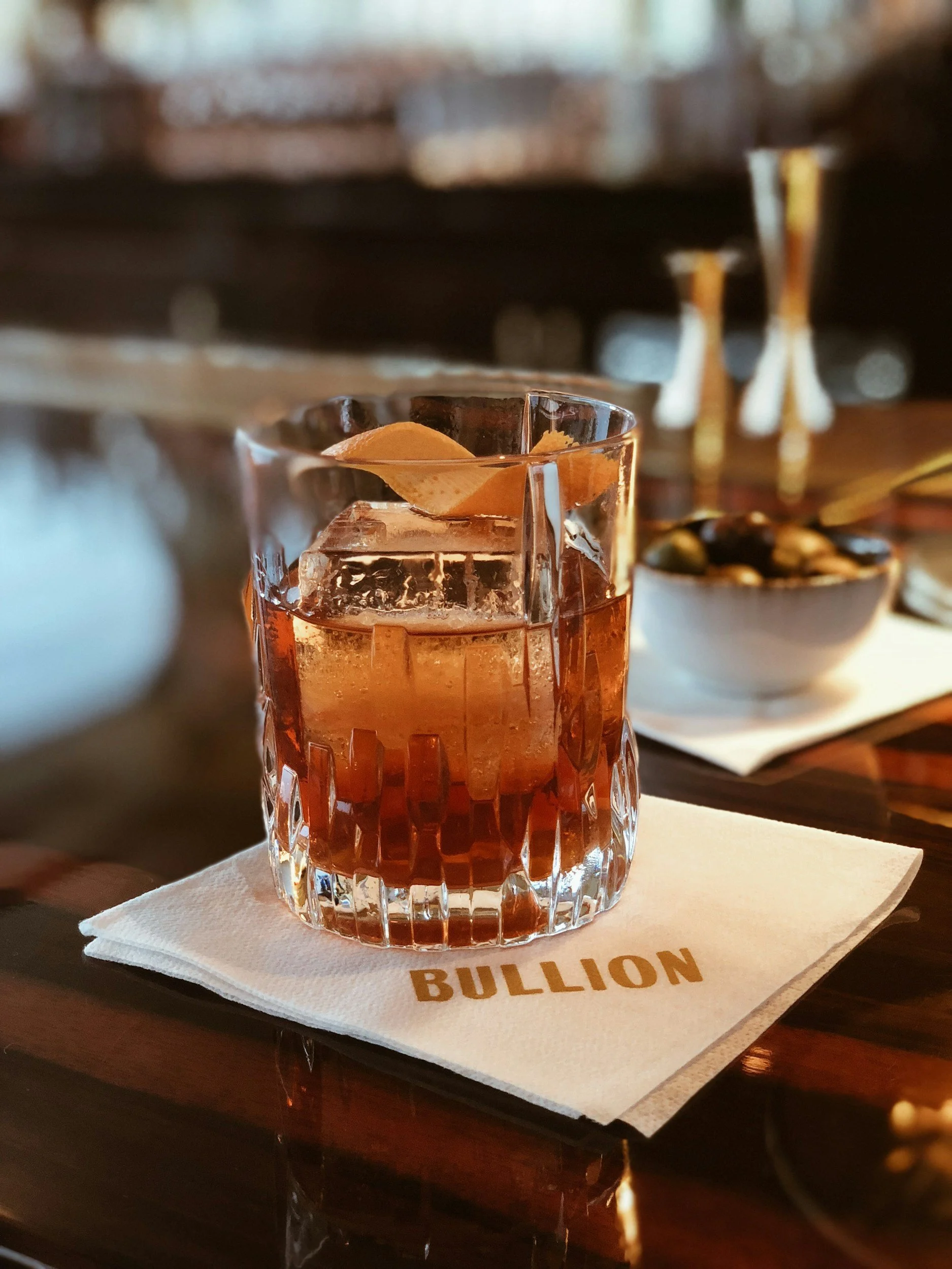 A glass of amber-colored cocktail with ice cubes and lemon peel garnish on a white napkin with the word 'BULLION' printed on it, set on a wooden table.