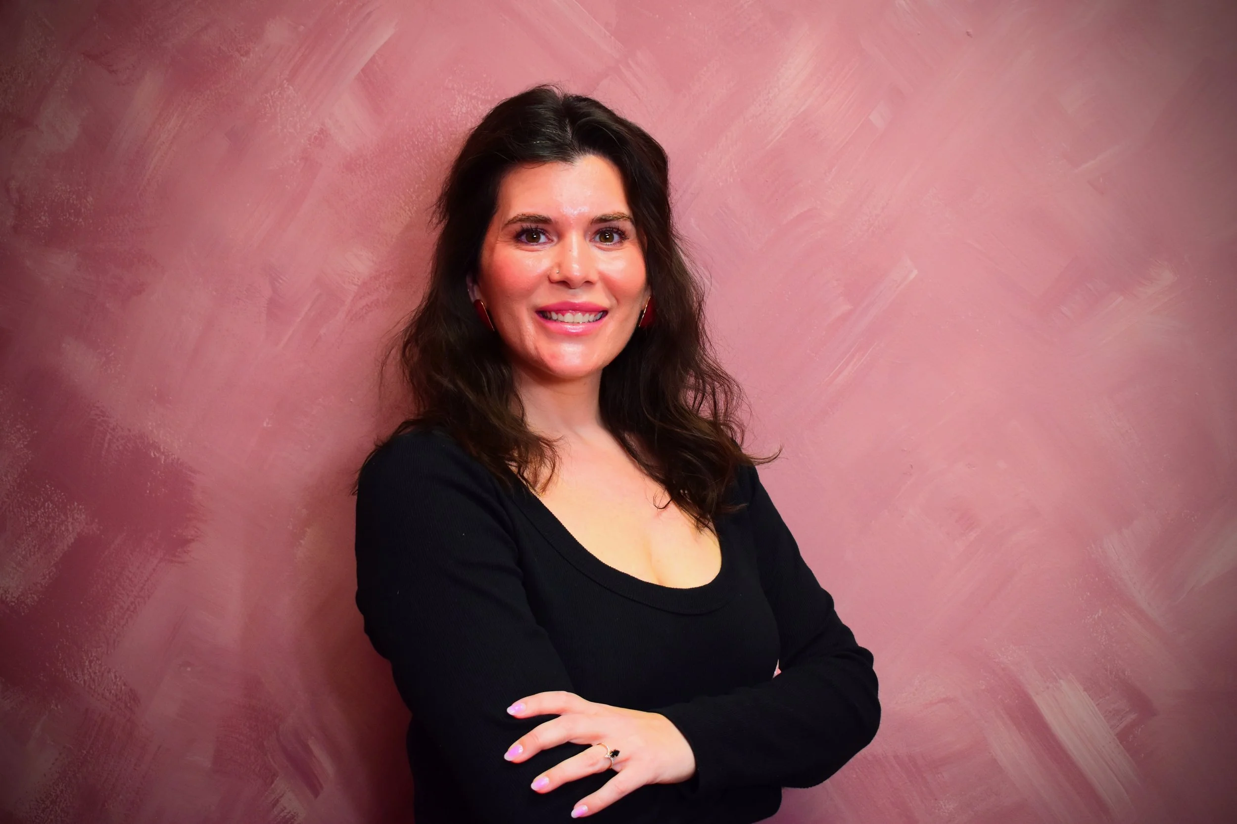 A woman with dark brown hair, wearing a black top, stands with her arms crossed in front of a pink textured wall, smiling at the camera. HWY Space #u02A- Sola Salons, 2901 South Capital of, Ste. 22, Austin, TX 78746, United States