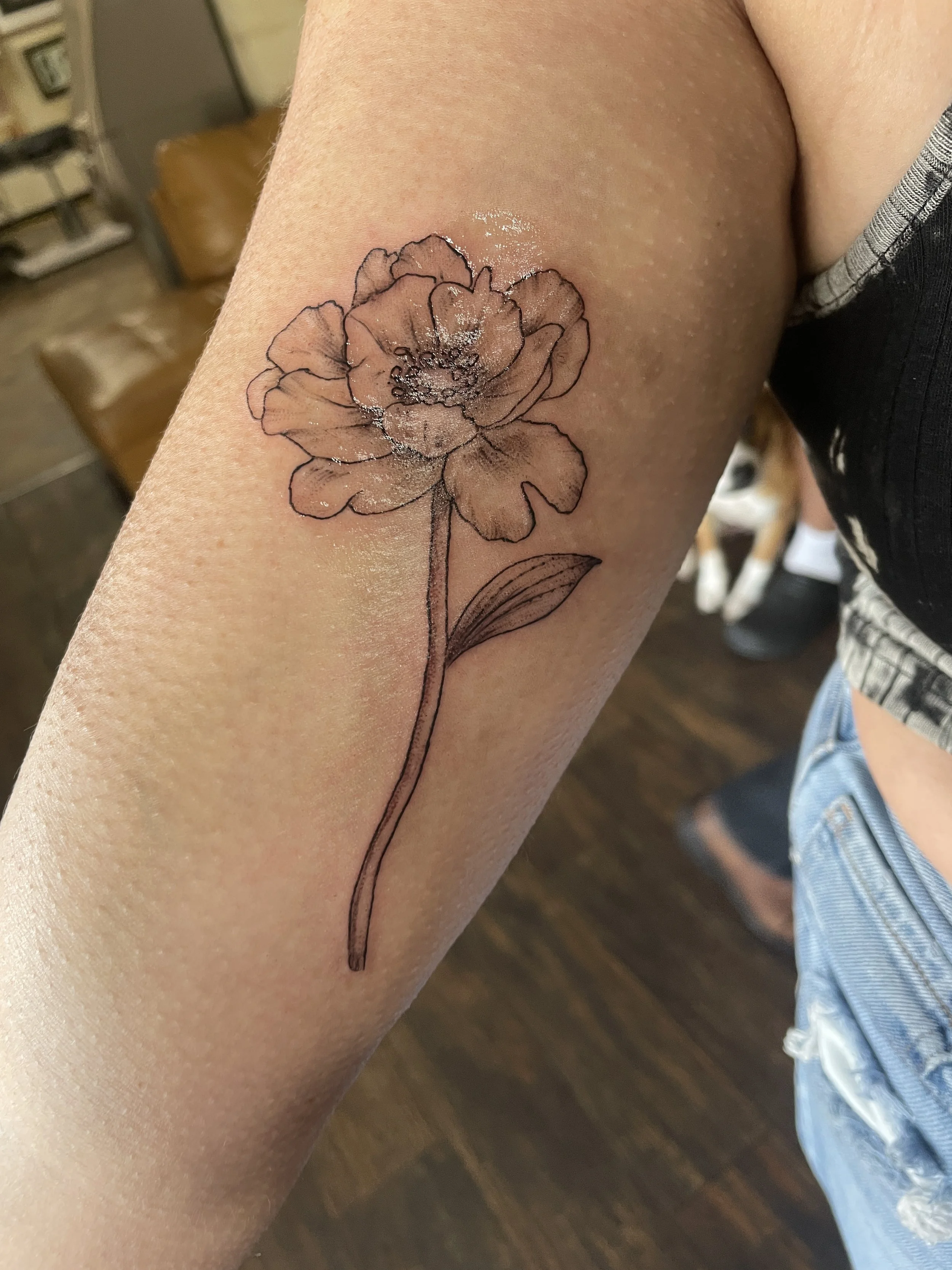 A person's arm with a flower tattoo on the upper arm. The tattoo is a delicate outline of a blooming flower, with some shiny white highlights.