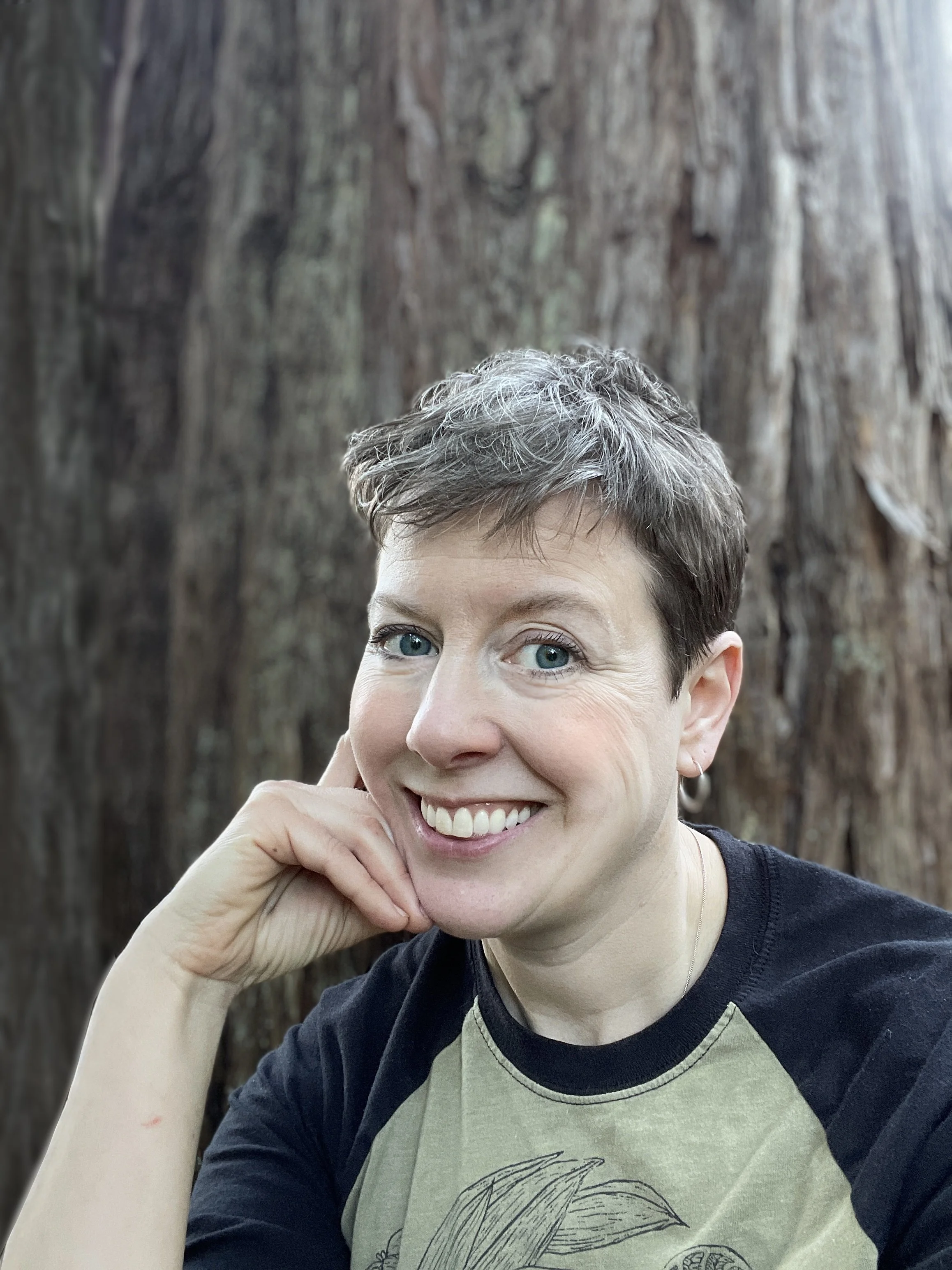 Close-up of a smiling woman with short hair, blue eyes, and earrings, resting her chin on her left hand, standing in front of a large tree trunk.