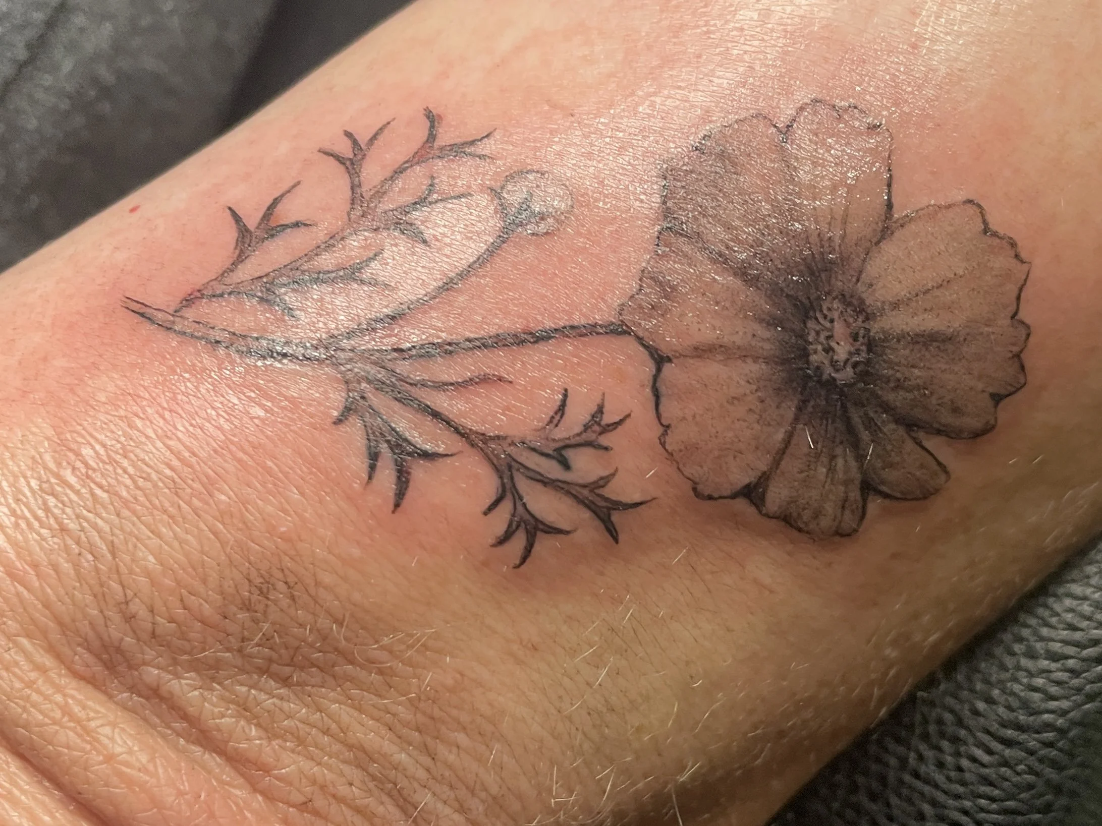 A tattoo of a fine line gray shaded outline of a botanical flower with a stem and leaves on person's skin. female tattoo artist Wednesday Kirwan