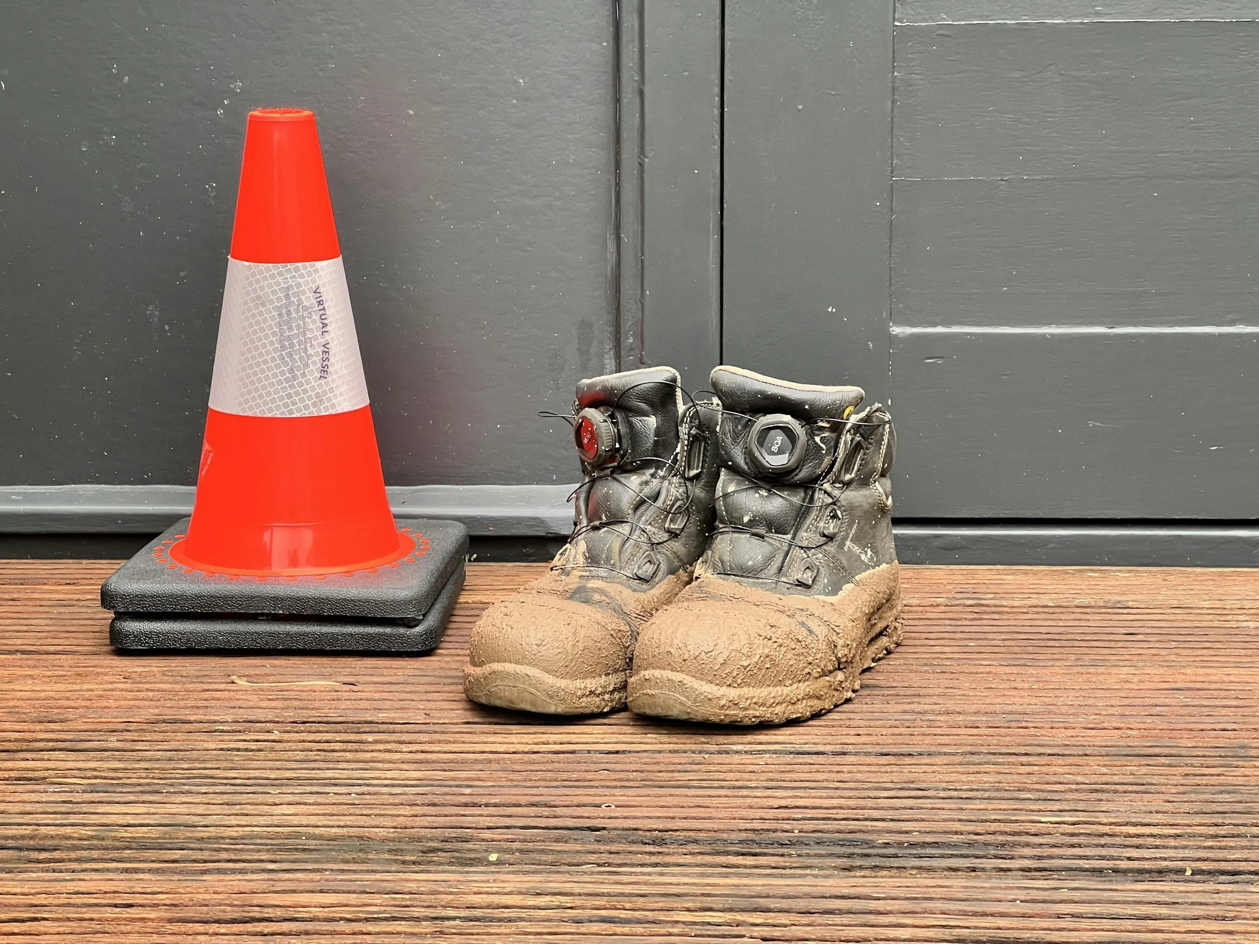 Pair of black and brown work boots covered in dirt placed on a wooden surface next to an orange traffic cone.