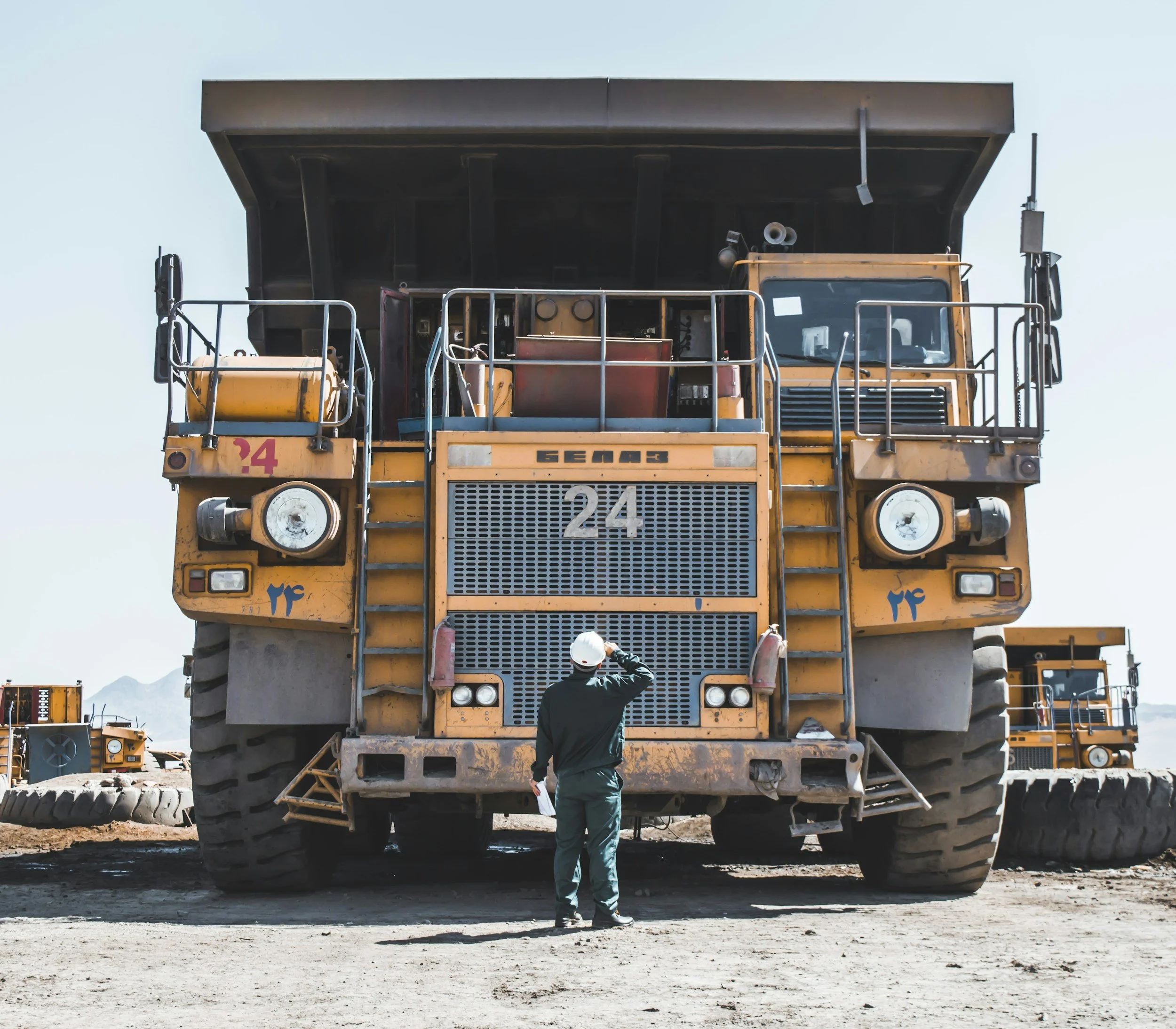 A construction worker in a hard hat and black jacket standing in front of a large yellow dump truck, inspecting the vehicle with one hand on his head. The truck is on a dirt construction site with other large construction vehicles in the background.